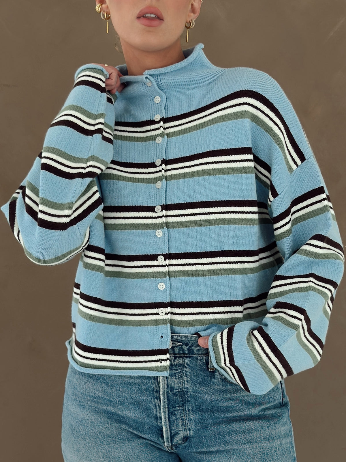 A person wearing a light blue knit button-front cardigan with horizontal brown, white, and olive green stripes, paired with blue jeans against a plain taupe background.