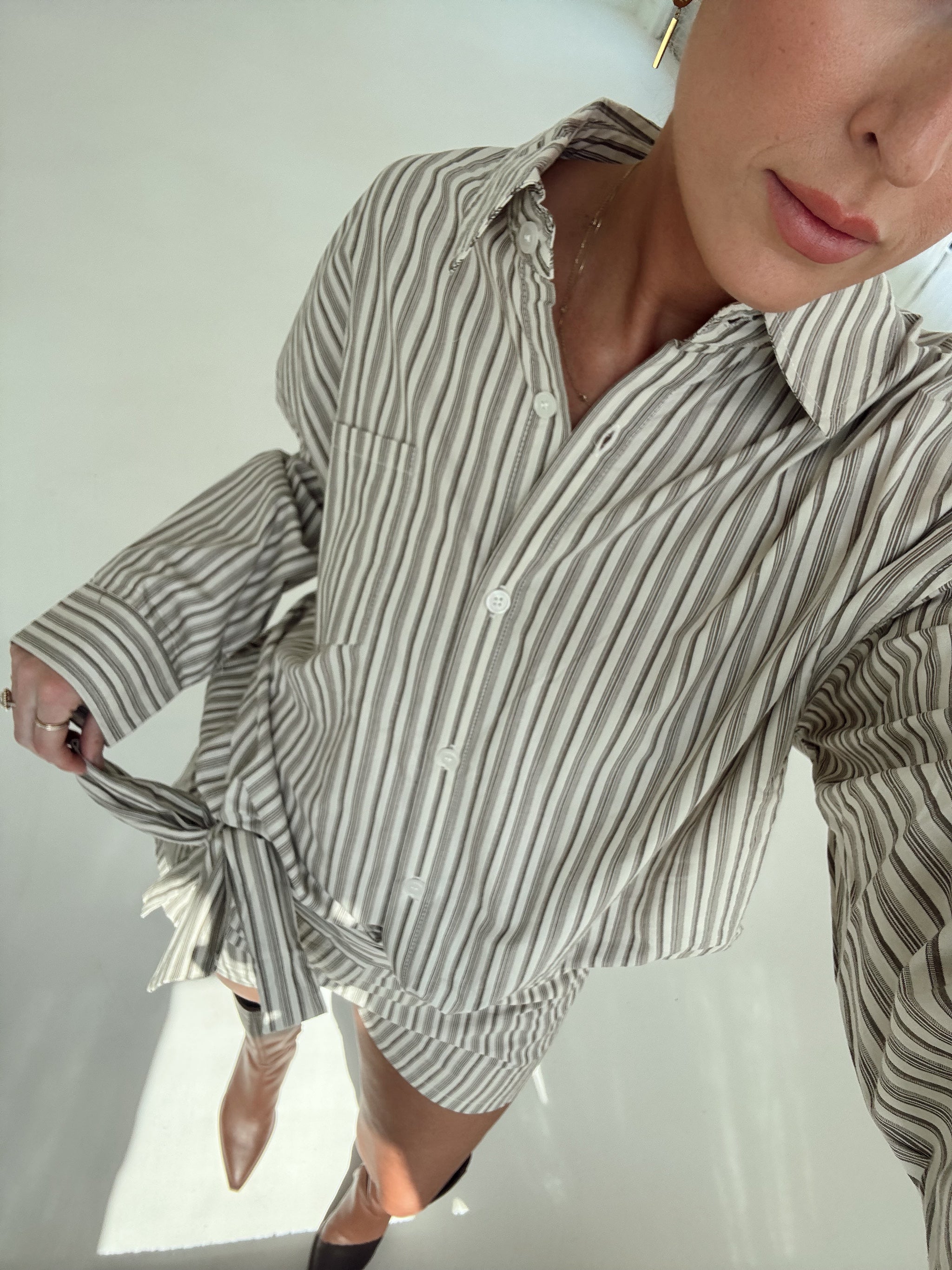 A woman stands in a bright, minimalist white room wearing a matching beige and white vertically striped button-down shirt and shorts set with a tie waist, paired with dark brown knee-high boots.