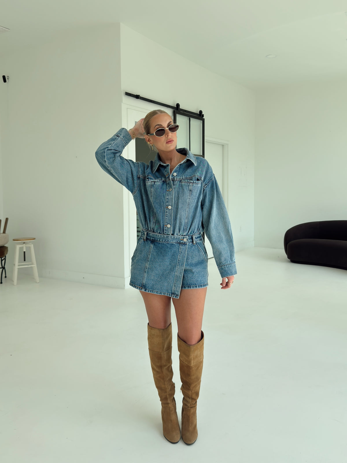 A woman modeling a blue denim romper, paired with brown suede over-the-knee boots, in a bright white studio setting.