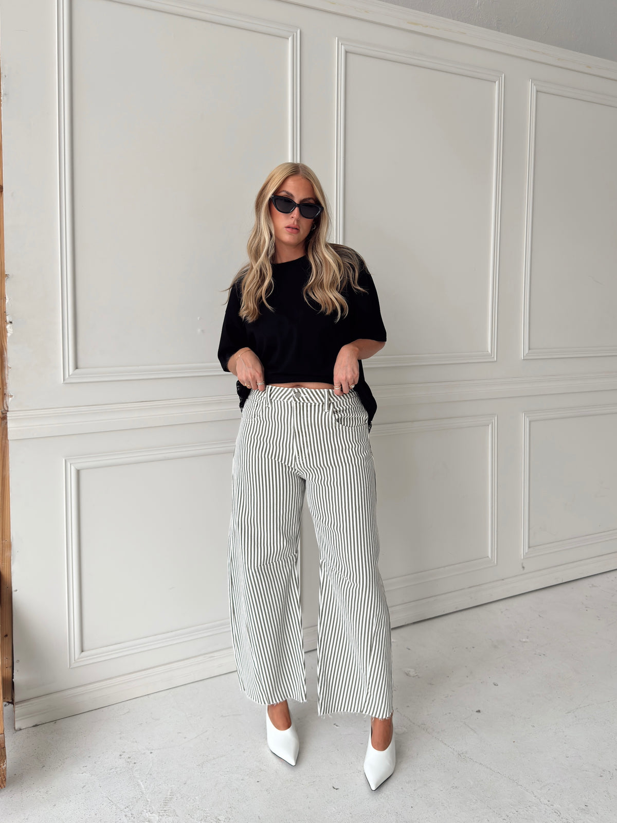 A woman modeling a pair of wide-leg barrel jeans with thin olive and white vertical stripes, paired with a black t-shirt, black sunglasses, and white pointed-toe heels, standing against a white paneled wall.
