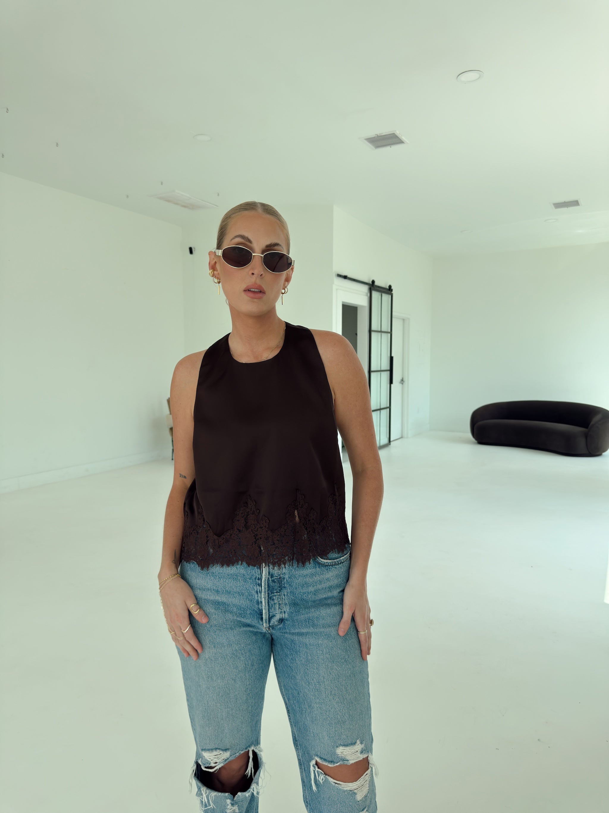 A woman wearing a brown sleeveless top with a frayed hem and light-wash distressed jeans, standing in a bright, minimalist white room.