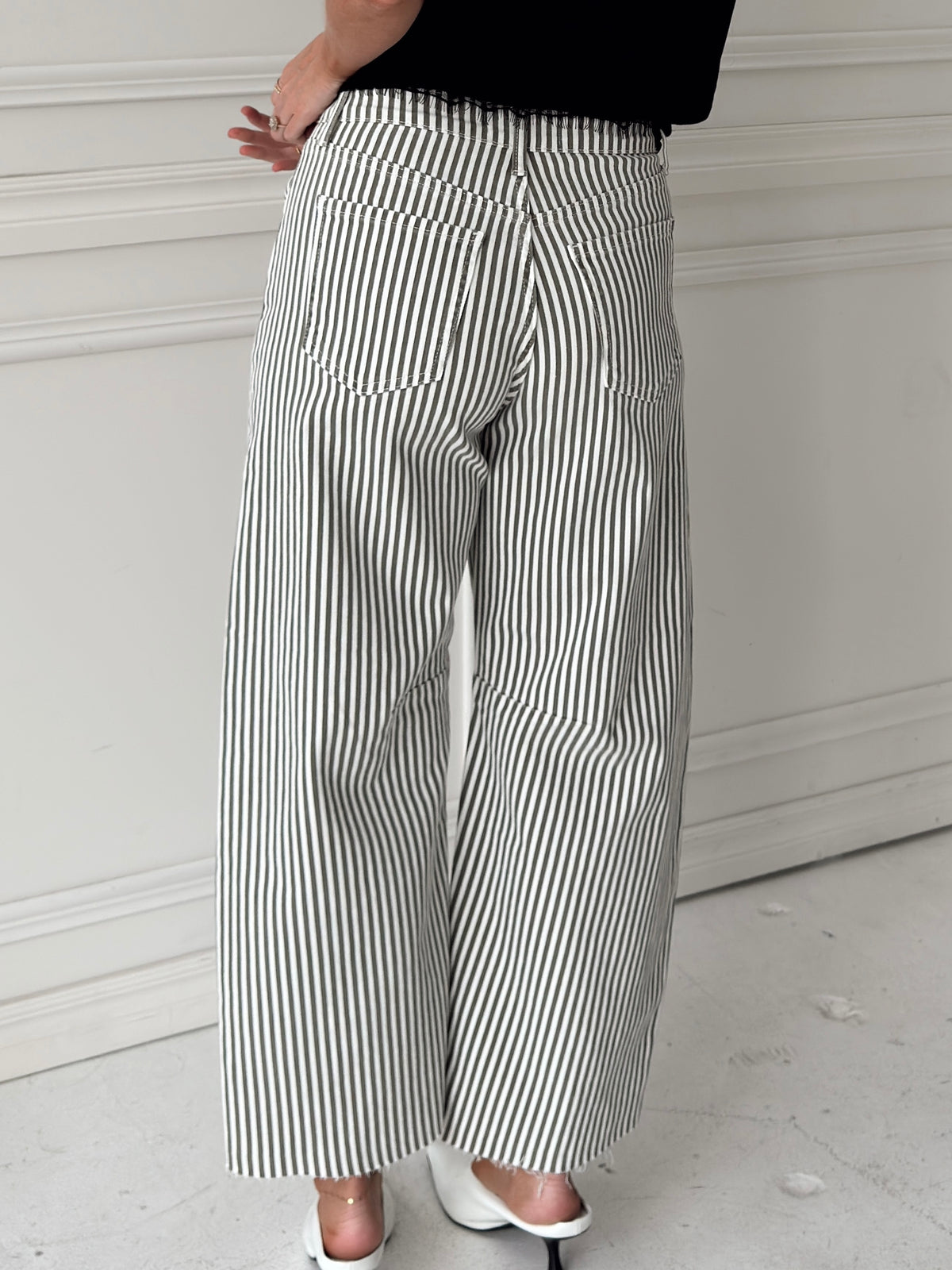 A person shown from the back wearing olive and white vertically striped wide-leg pants, a black top, and white heeled sandals, standing against a white paneled wall.