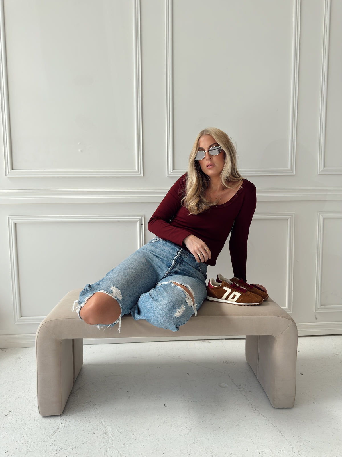 A woman wearing a burgundy long-sleeve top, ripped light-wash blue jeans, and brown sneakers sits on a modern beige bench against a white paneled wall.