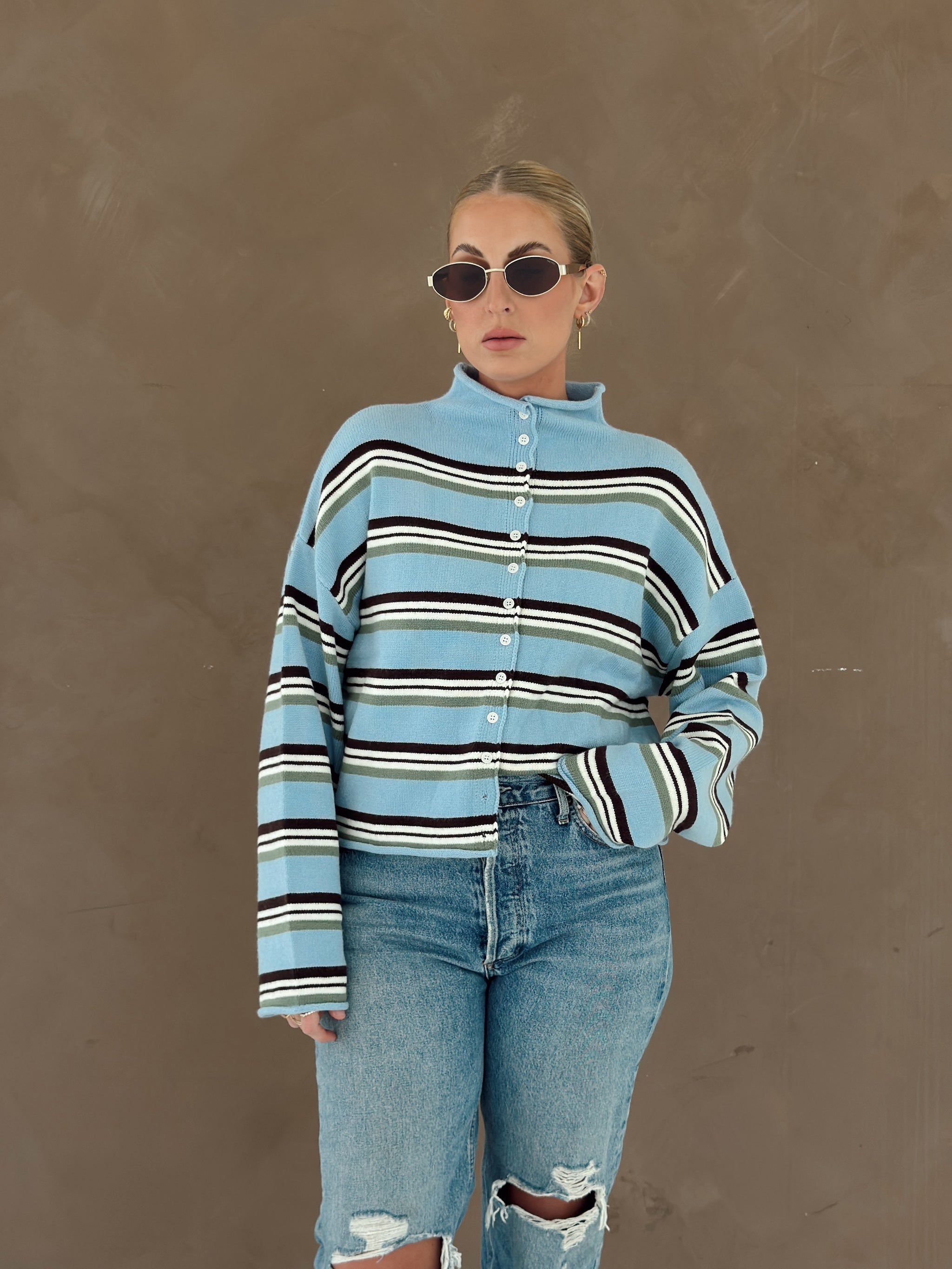 A person wearing a light blue knit cardigan with horizontal brown, white, and olive green stripes, paired with light-wash jeans.