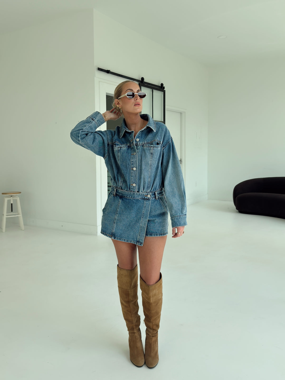 A woman modeling a blue denim romper, paired with brown suede over-the-knee boots, in a bright white studio setting.