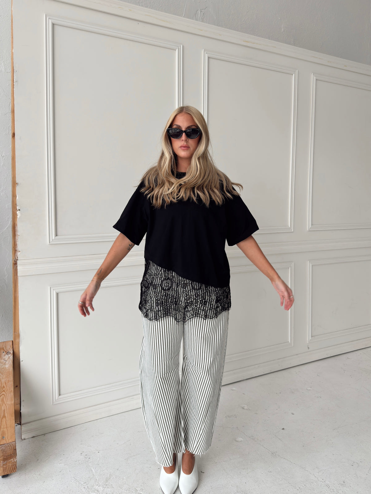 A woman modeling a black short-sleeve top with a sheer black lace hem, paired with white and olive vertically striped wide-leg pants and white pointed shoes, standing in front of a white paneled wall.