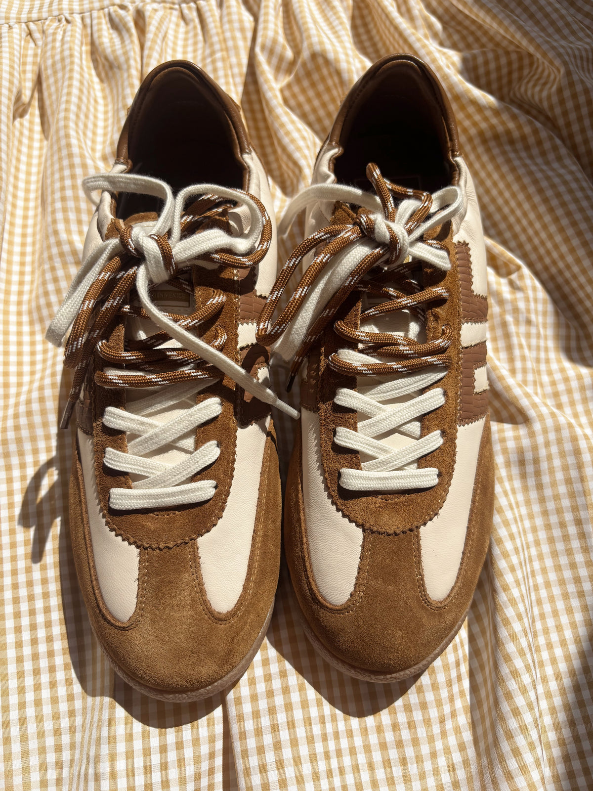 A pair of retro-style low-top sneakers in brown suede and off-white leather, featuring chunky white laces and three stripes on the side, laid flat on a beige and white gingham fabric background.