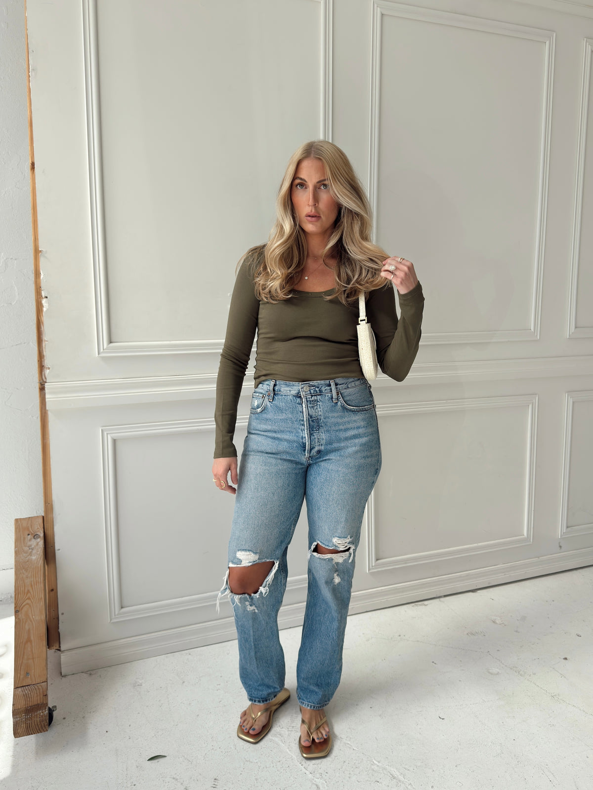 A woman stands indoors wearing a fitted olive green long-sleeve top, light blue ripped jeans, and sandals, holding a cream shoulder bag. The background is a white paneled wall.