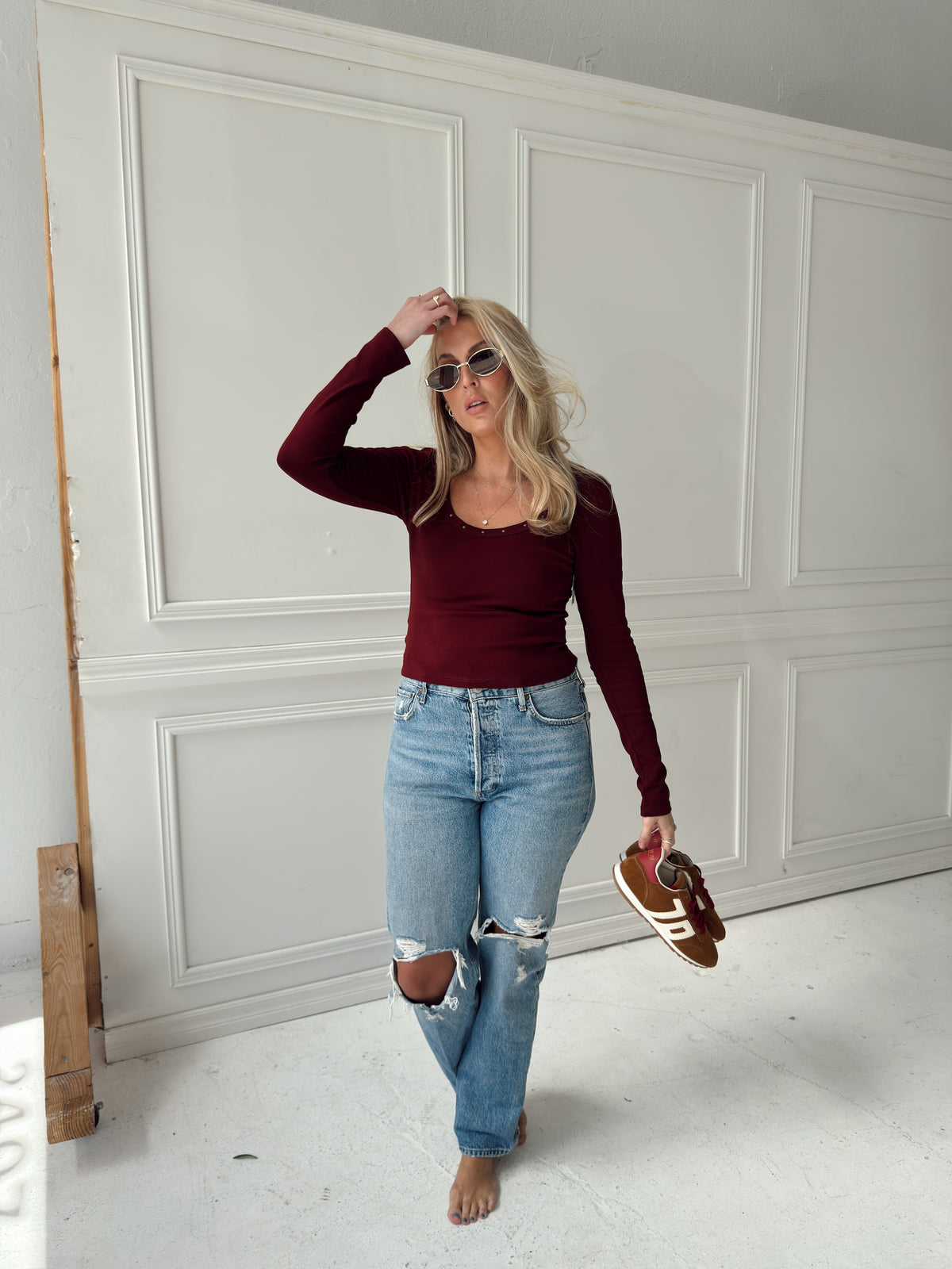 A woman stands in a white paneled room wearing a fitted burgundy long-sleeve top, light-wash distressed blue jeans, and brown sneakers.