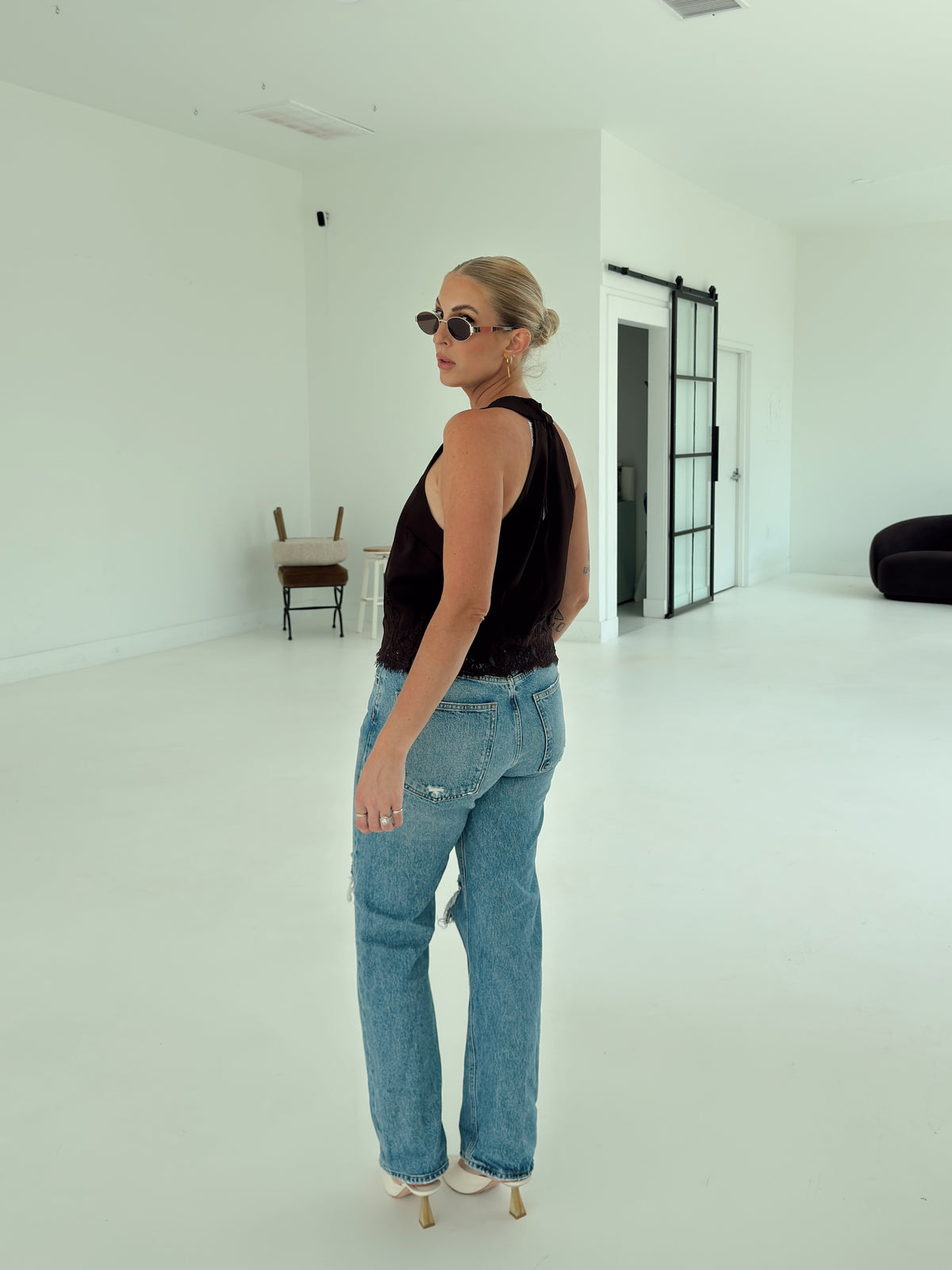 A woman wearing a brown sleeveless top and light blue straight-leg jeans stands in a bright, minimalist white studio space, looking over her shoulder.