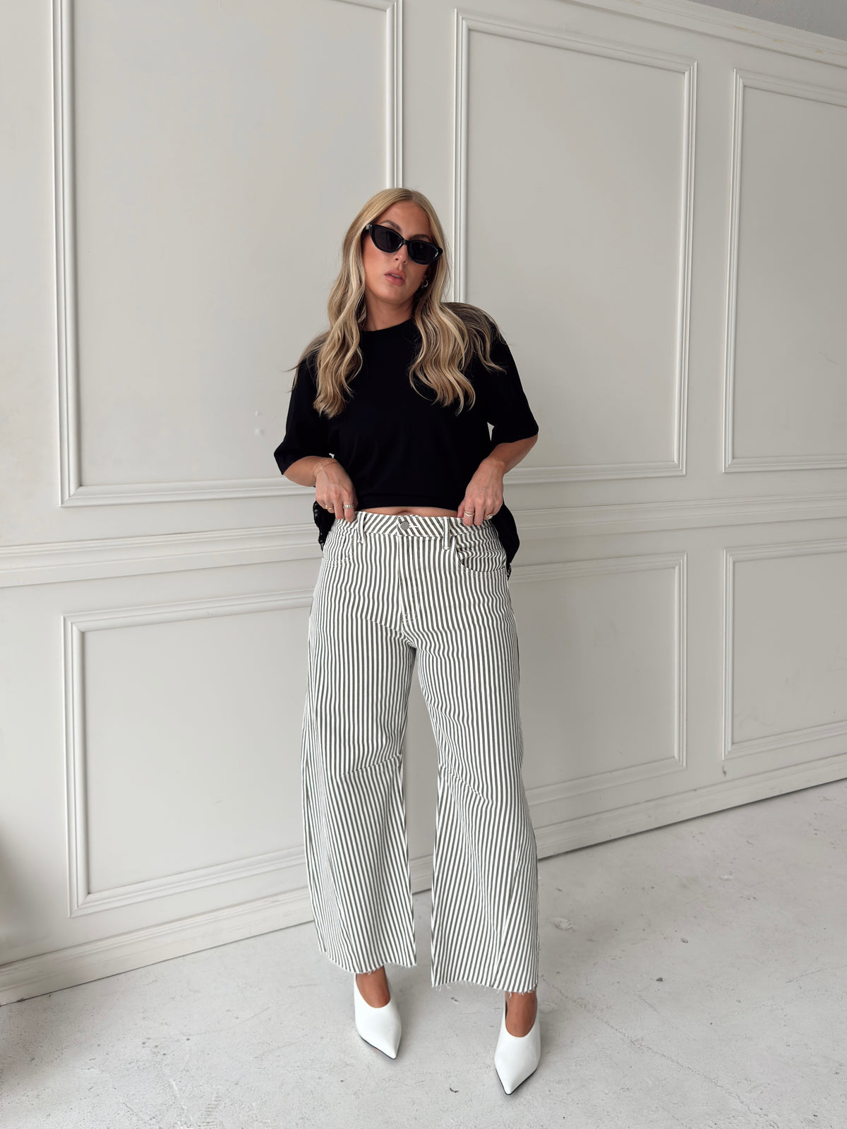 A woman modeling a pair of wide-leg barrel jeans with thin olive and white vertical stripes, paired with a black t-shirt, black sunglasses, and white pointed-toe heels, standing against a white paneled wall.