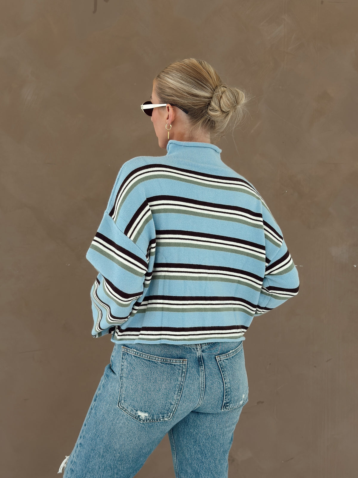 A woman shown from behind wearing a light blue striped mock-neck sweater with brown, white, and green stripes, paired with light-wash blue jeans, against a textured brown wall.