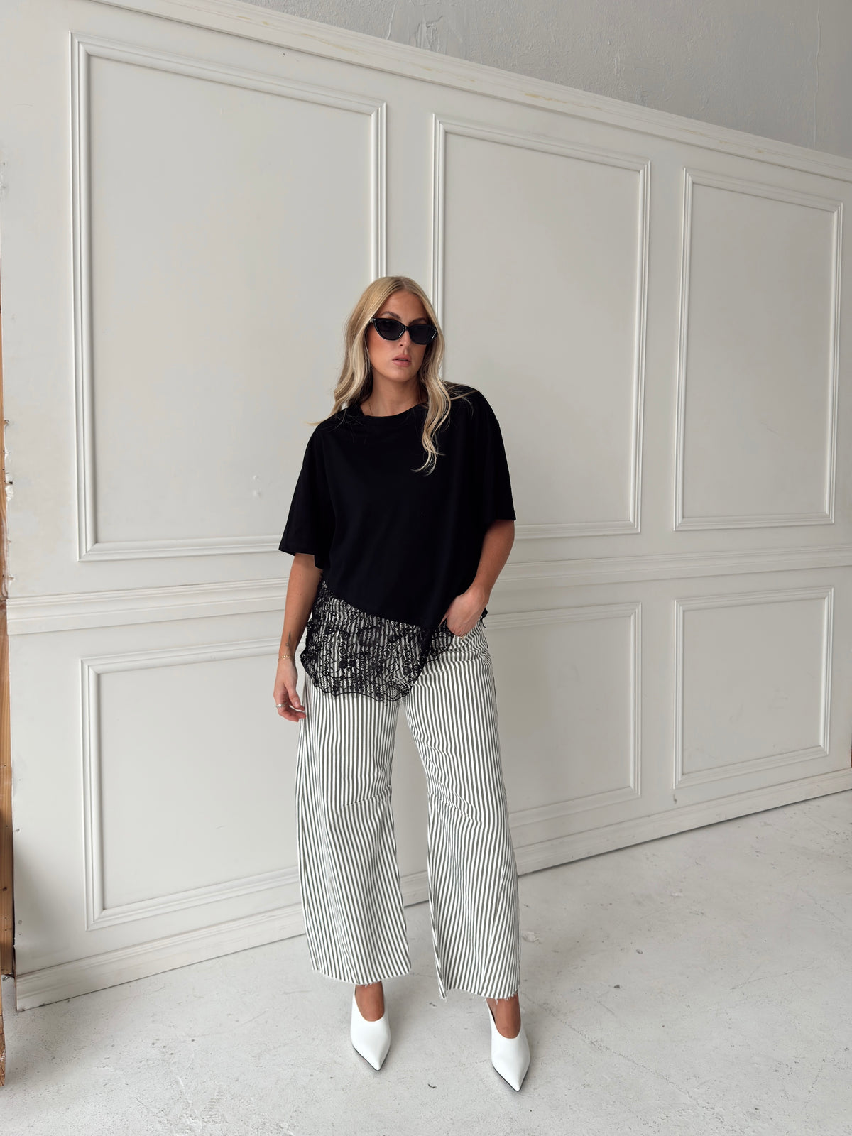 A woman modeling a black asymmetrical top with a sheer black lace hem, paired with white and olive vertically striped pants, standing in front of a white paneled studio backdrop.