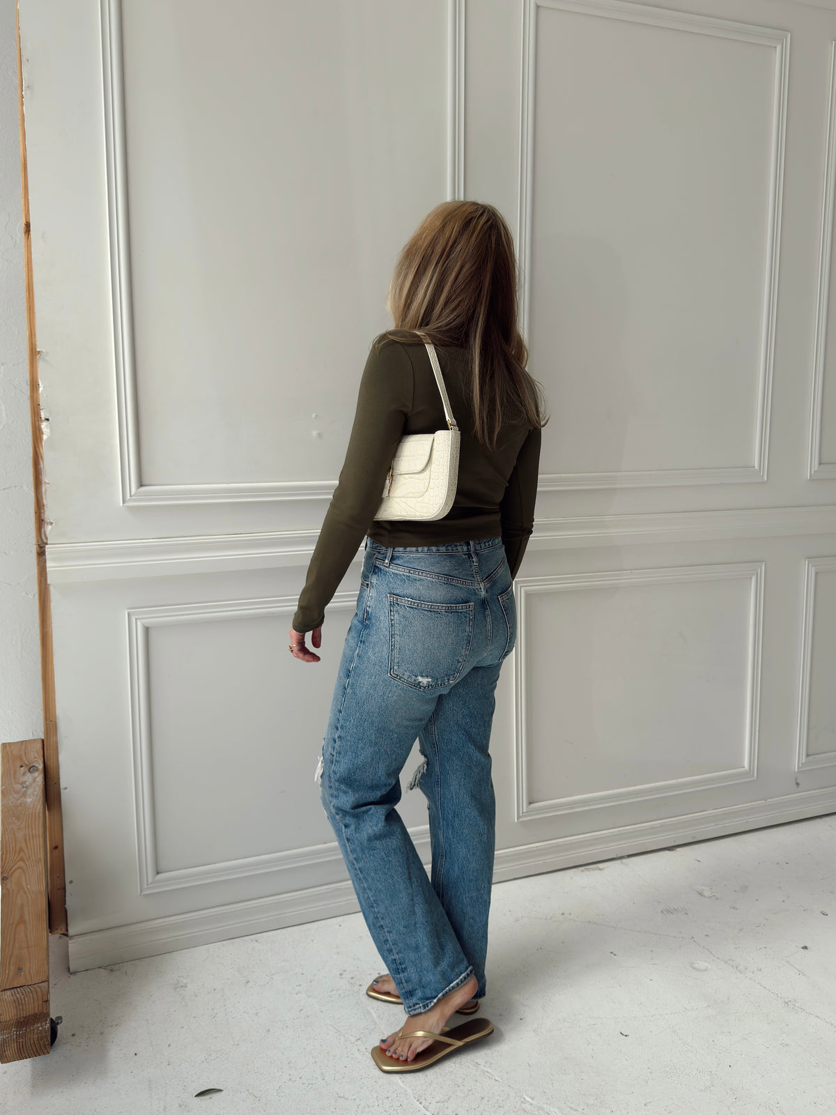 A woman is shown from behind wearing a dark olive green long-sleeve top, blue straight-leg jeans, and sandals, carrying a small white shoulder bag against a white paneled wall.