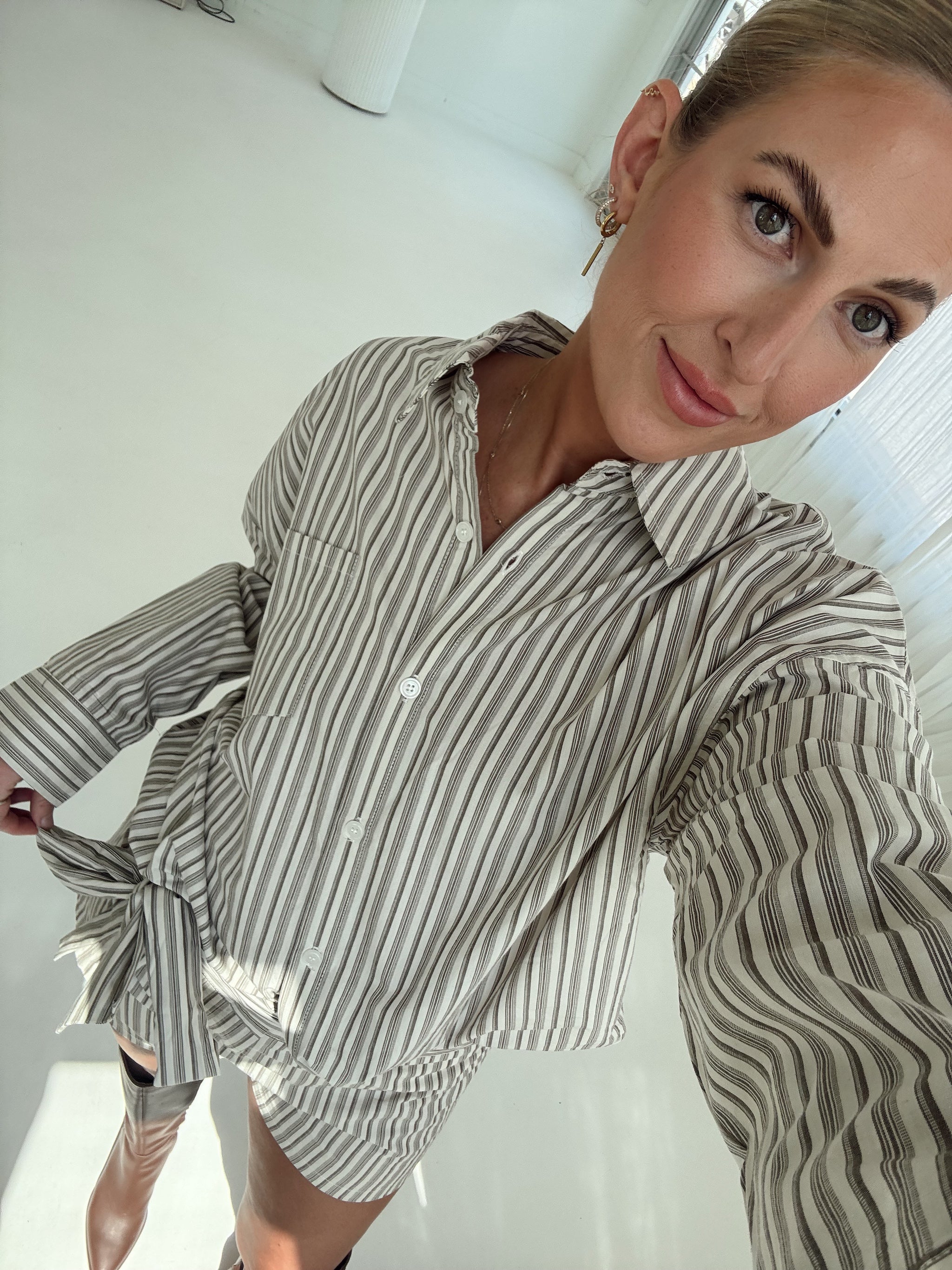 A woman wearing a beige and dark brown vertically striped button-down shirt, taking a mirror selfie in a bright, minimalist room.