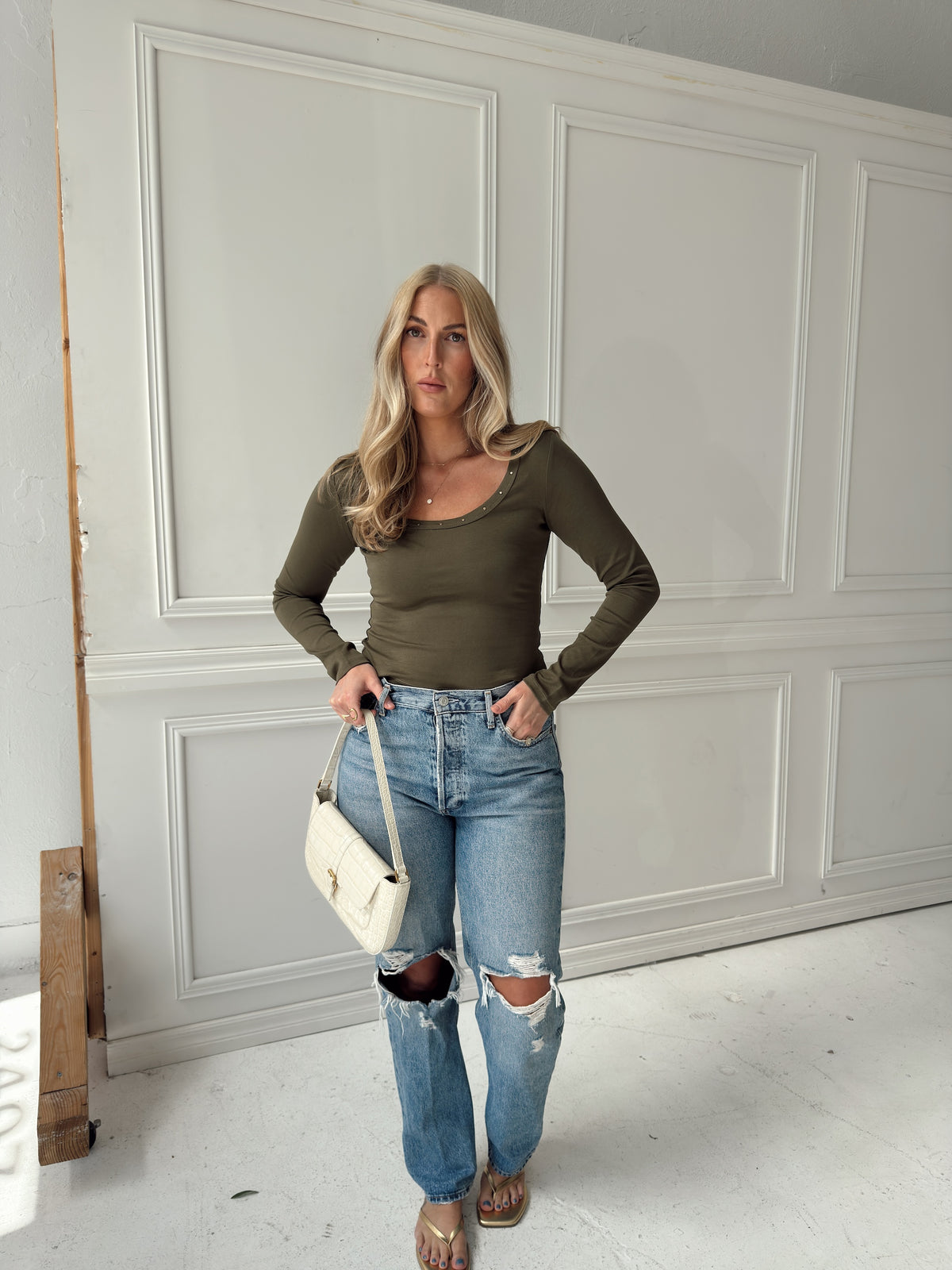 A woman stands in a studio wearing a fitted olive green long-sleeve scoop-neck top, light-wash distressed blue jeans with ripped knees, and carrying a small cream shoulder bag.