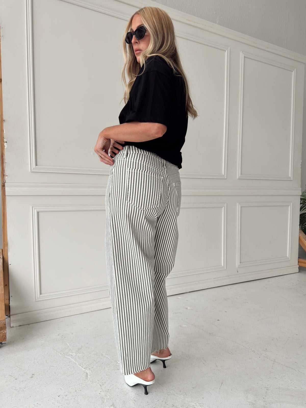 A woman modeling a pair of high-waisted, wide-leg jeans with thin olive vertical stripes on an off-white base. She is wearing a black short-sleeve top and white heeled sandals, standing in front of a white paneled wall.