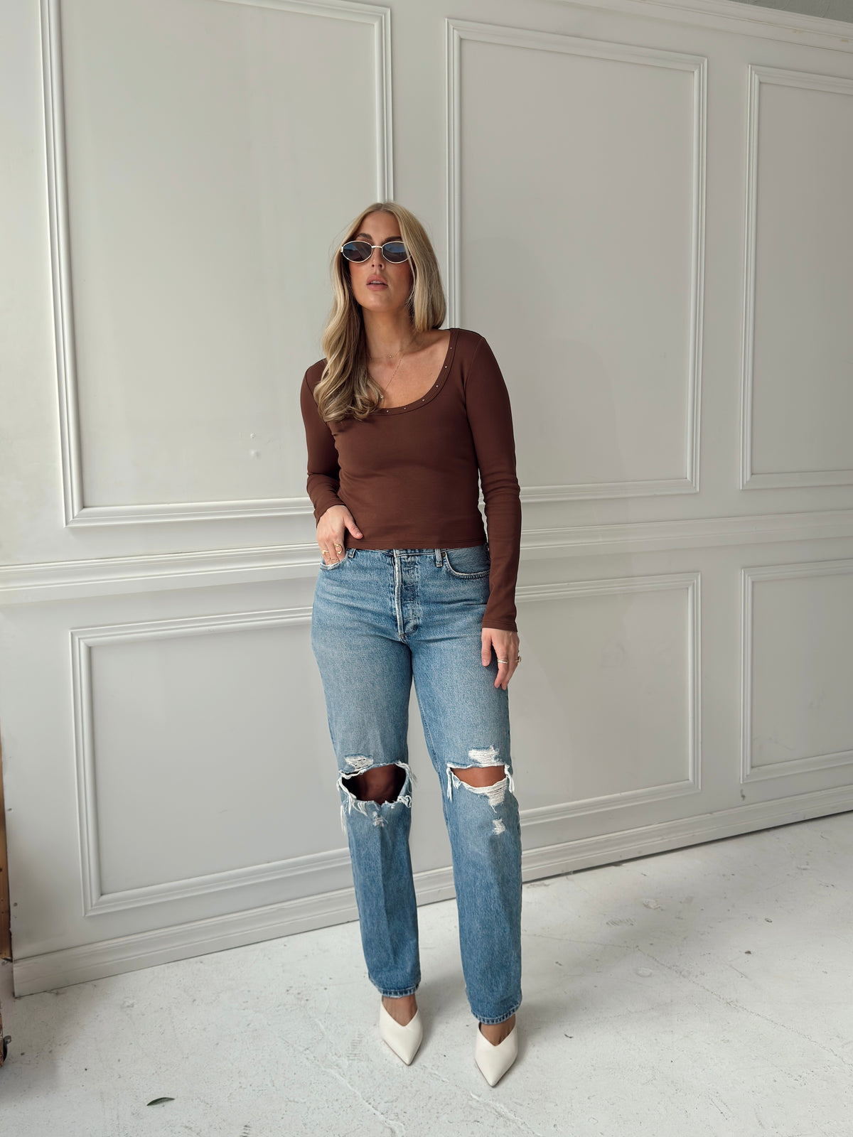 A woman modeling a brown long-sleeve scoop-neck top and light-wash distressed blue jeans with ripped knees, paired with white pointed-toe heels, standing against a white paneled wall.