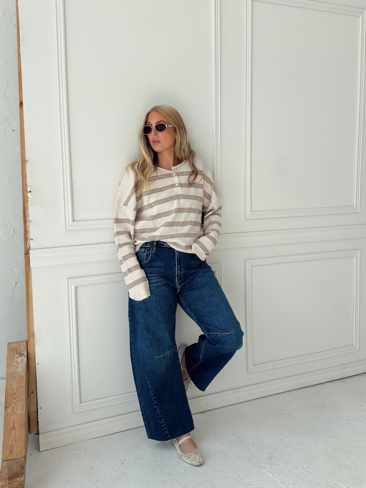 A woman stands in front of a white paneled wall wearing a beige, cream, and light blue horizontally striped long-sleeve knit top, wide-leg blue jeans, and light-colored flats.