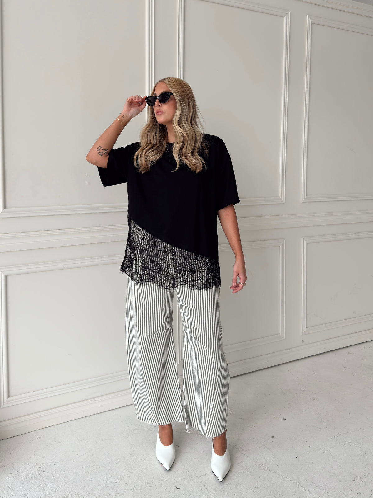 A woman modeling a black asymmetrical top with a sheer black lace hem, paired with white and olive vertically striped pants, standing in front of a white paneled studio backdrop.