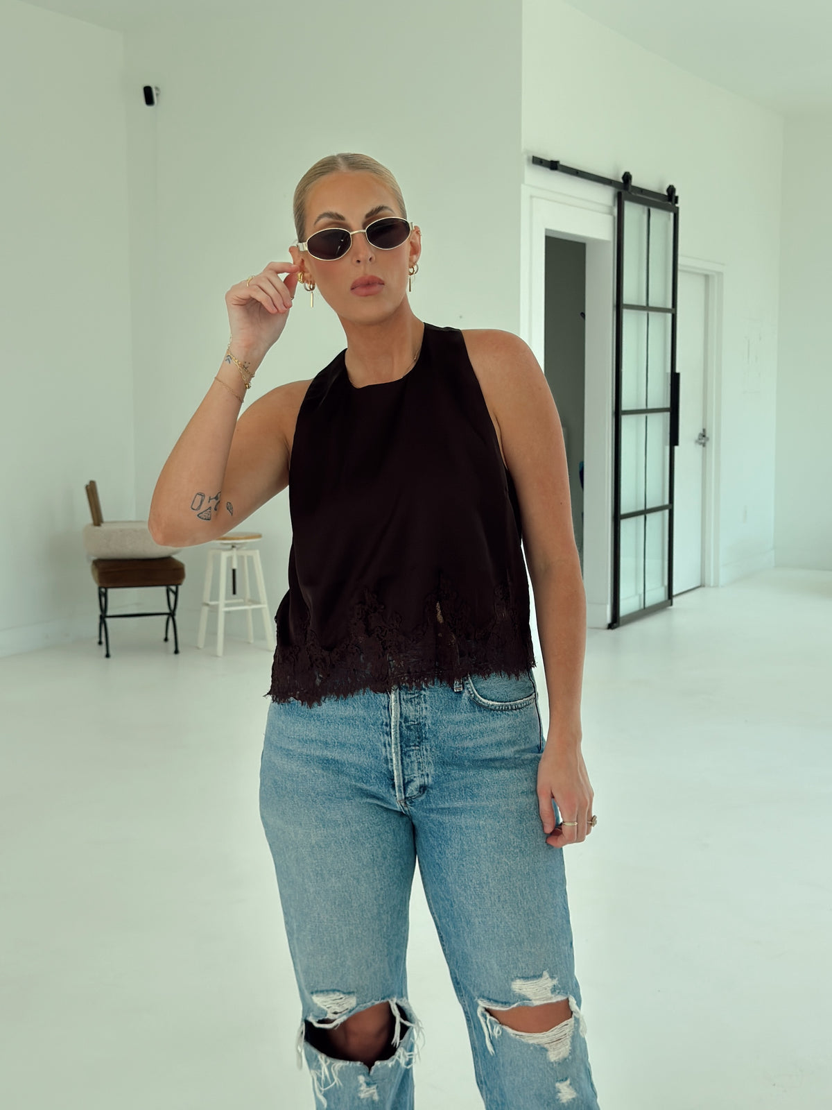 A woman posing in a minimalist white studio setting, wearing a brown sleeveless top, light-wash distressed blue jeans, and white pointed-toe heels.