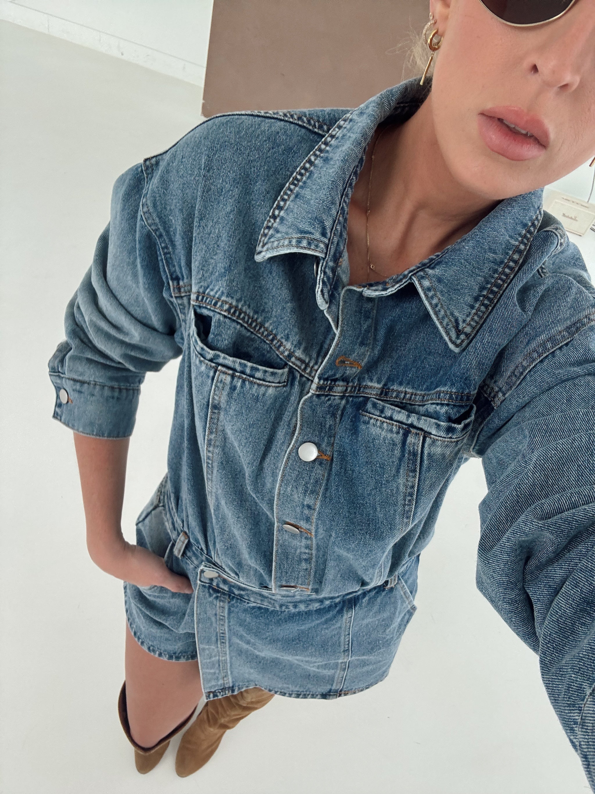 A woman wearing a light blue denim romper with a collar, button front, and chest pockets, paired with brown pointed-toe heels, photographed in a bright indoor setting.