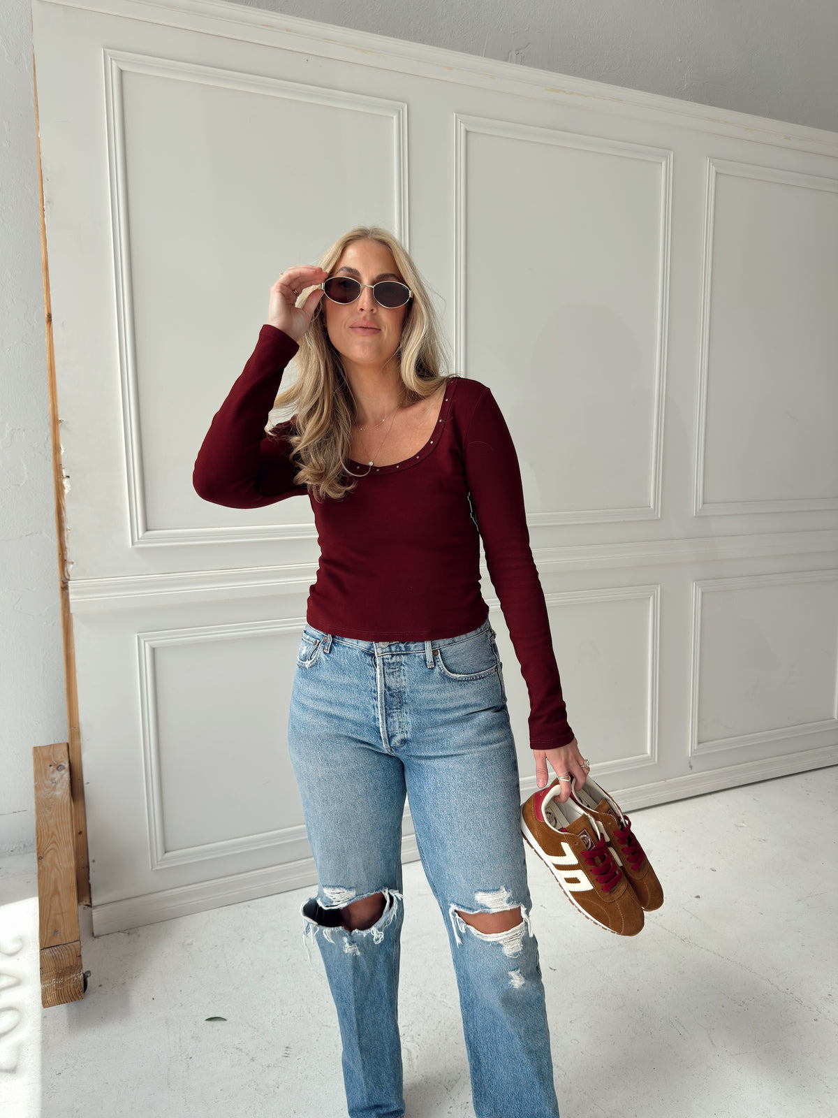 A woman stands indoors wearing a fitted burgundy long-sleeve top, light blue ripped jeans, and sunglasses, while holding a pair of brown and burgundy sneakers. She is posed in front of a white paneled wall.