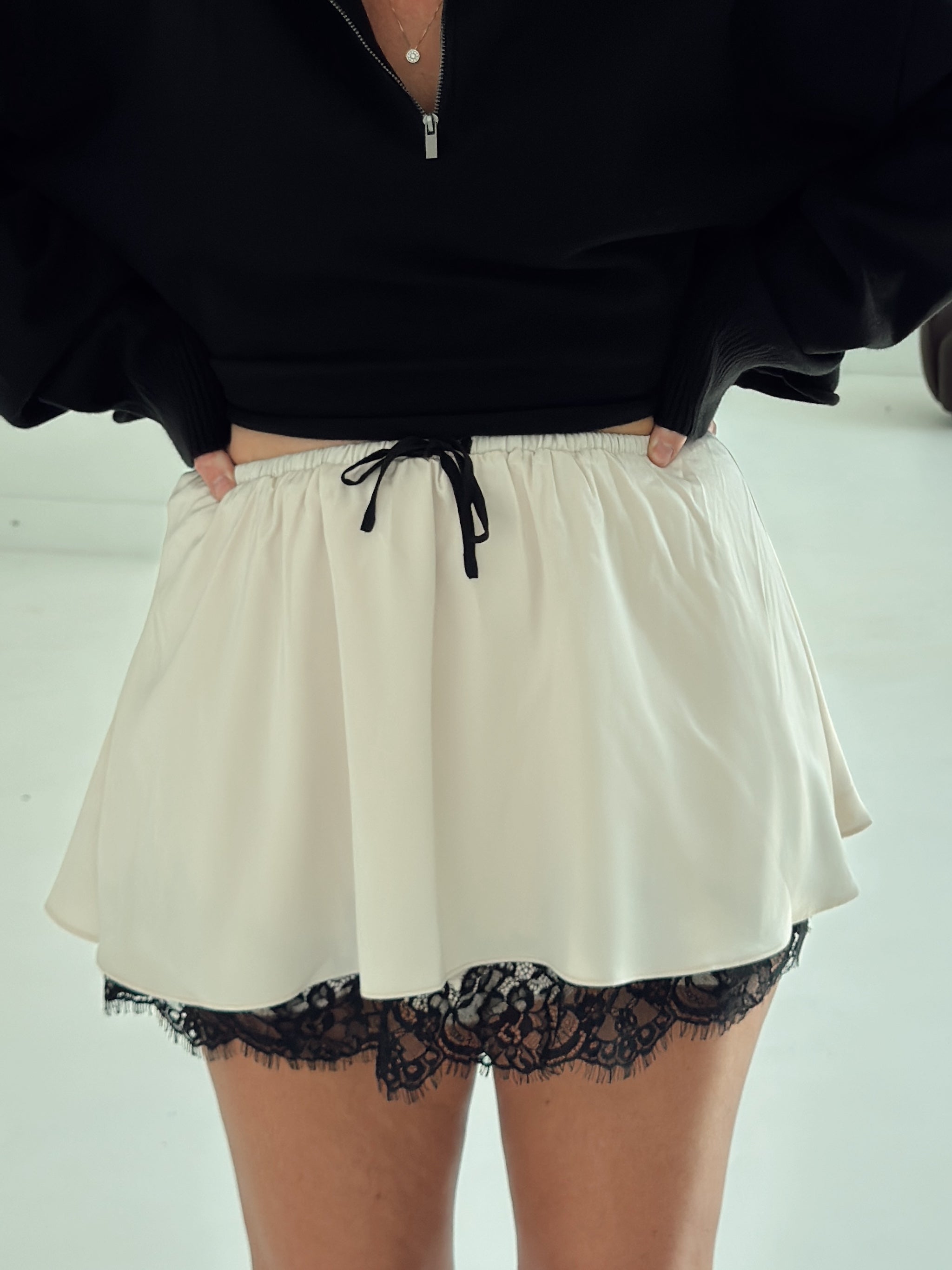 A person wearing a cream-colored mini skirt with a black lace hem and black drawstring, paired with a black zip-up top.