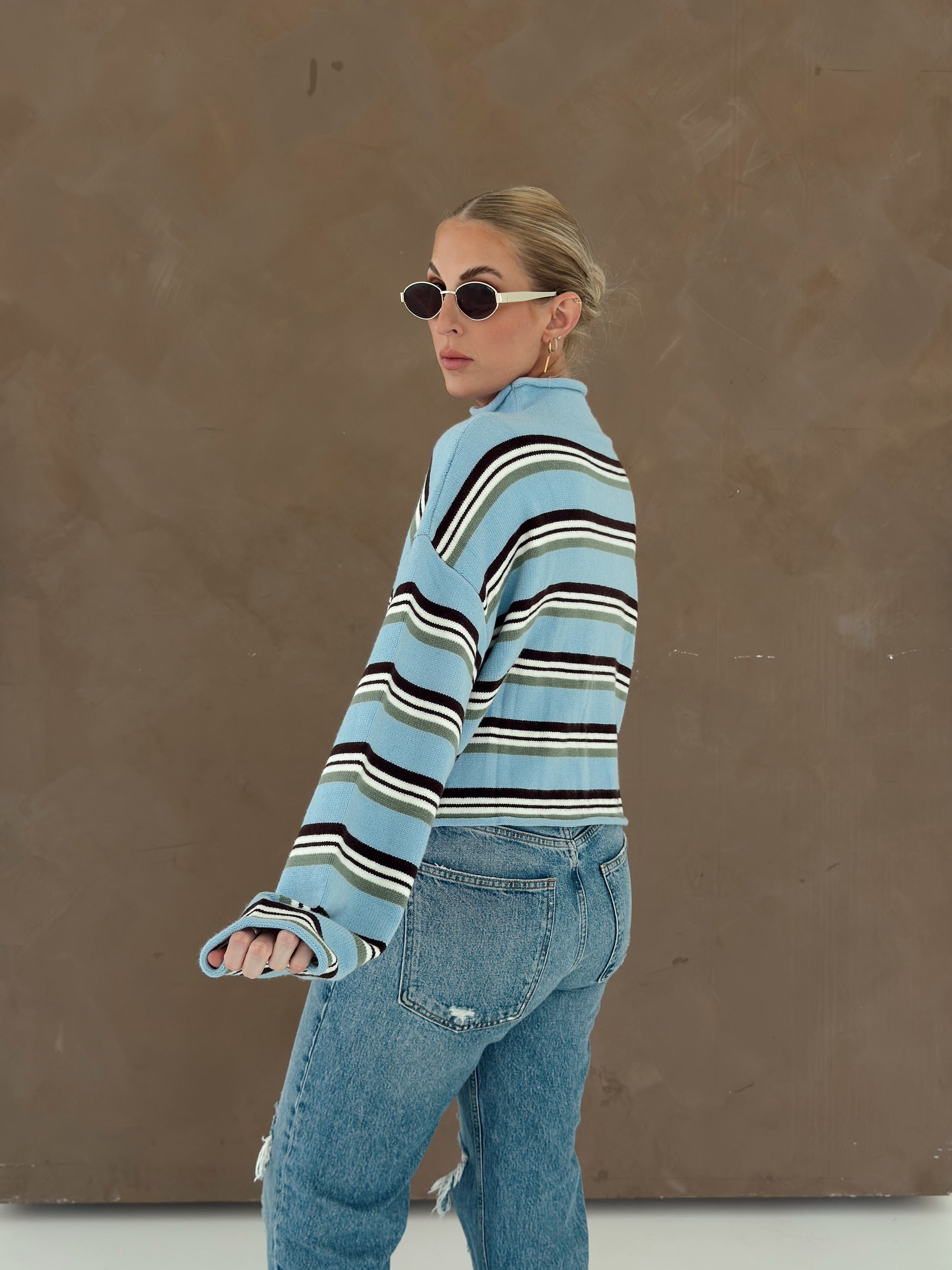 A woman wearing a light blue, brown, green, and white striped knit sweater and light-wash blue jeans, photographed from the back against a textured brown studio backdrop.