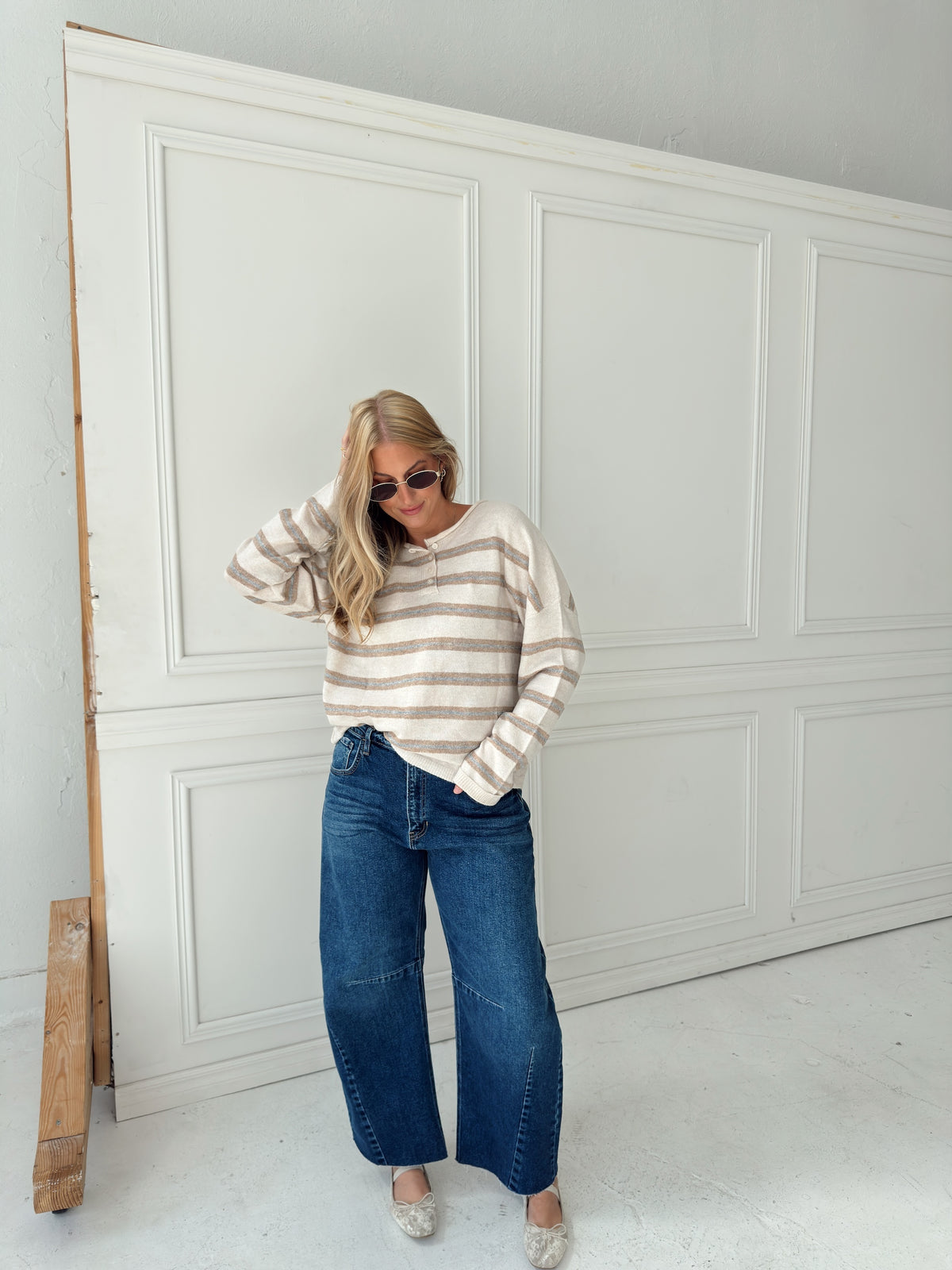 A woman stands in front of a white paneled wall wearing a beige, cream, and light blue horizontally striped long-sleeve knit top, wide-leg blue jeans, and light-colored flats.