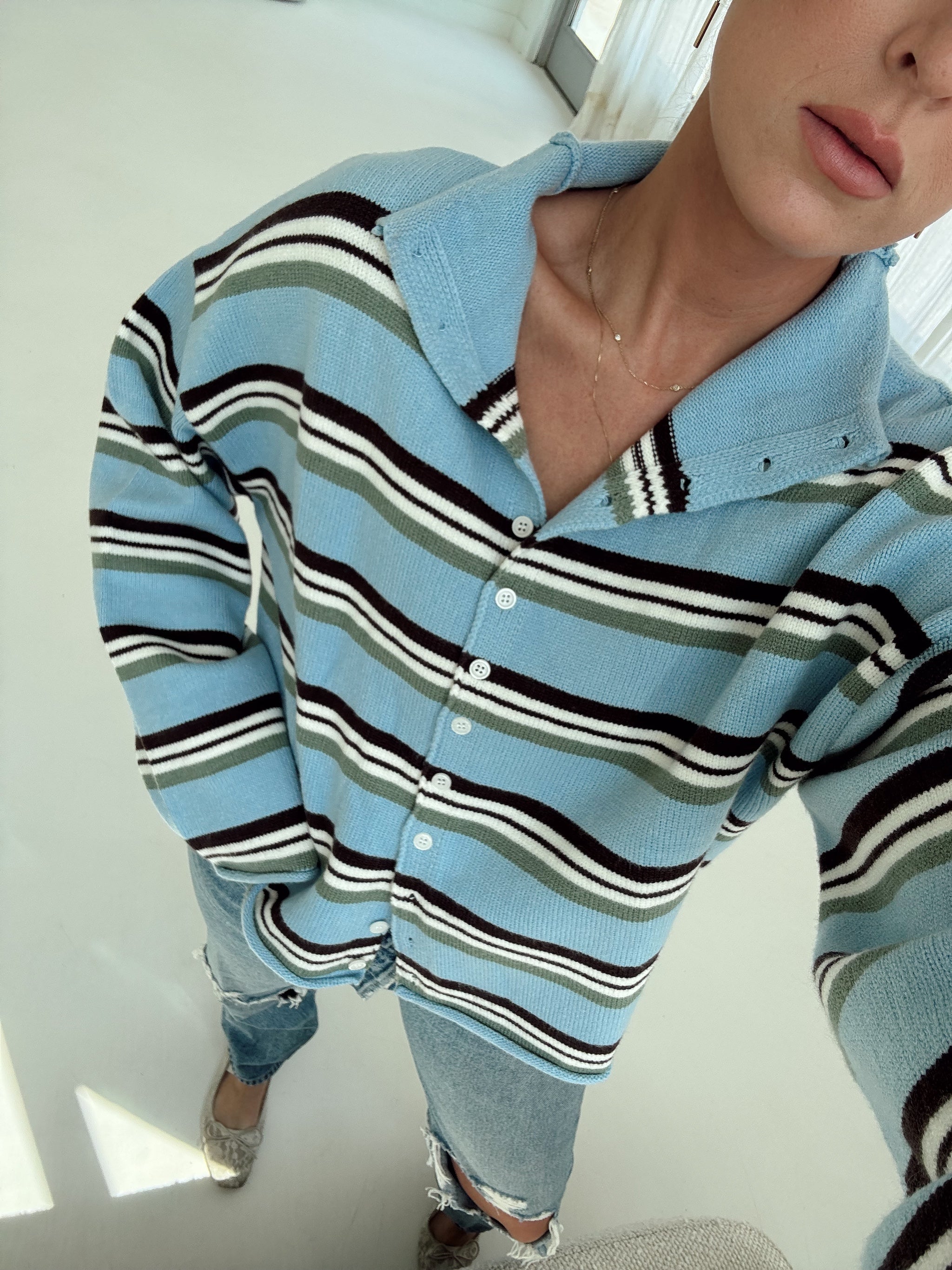 A person wearing a light blue knit cardigan with horizontal brown, white, and olive green stripes, paired with light-wash jeans and lace flats.