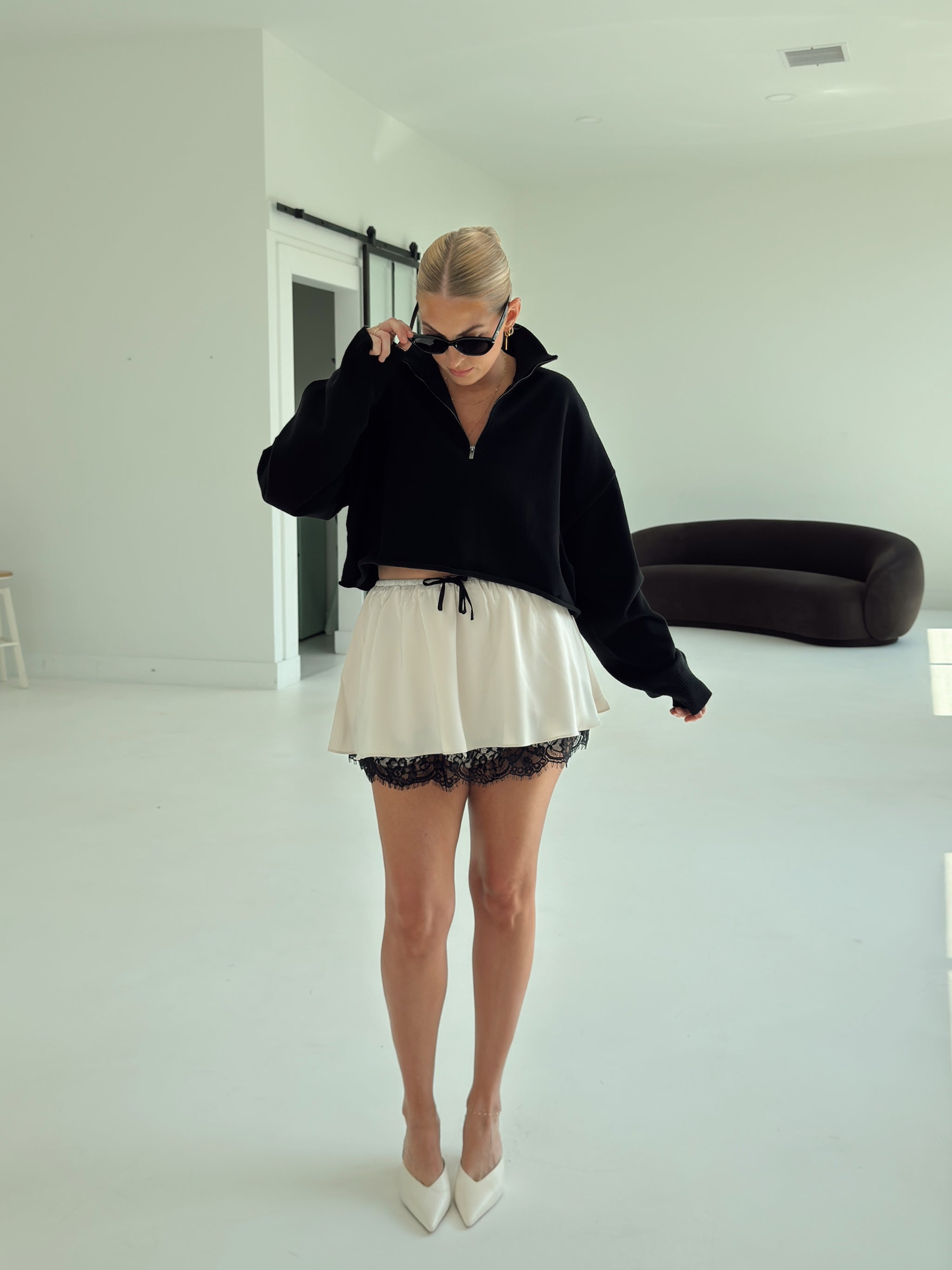 A woman stands in a bright white room wearing a black cropped zip-up hoodie, a cream mini skirt with black lace trim, and white pointed-toe heels.