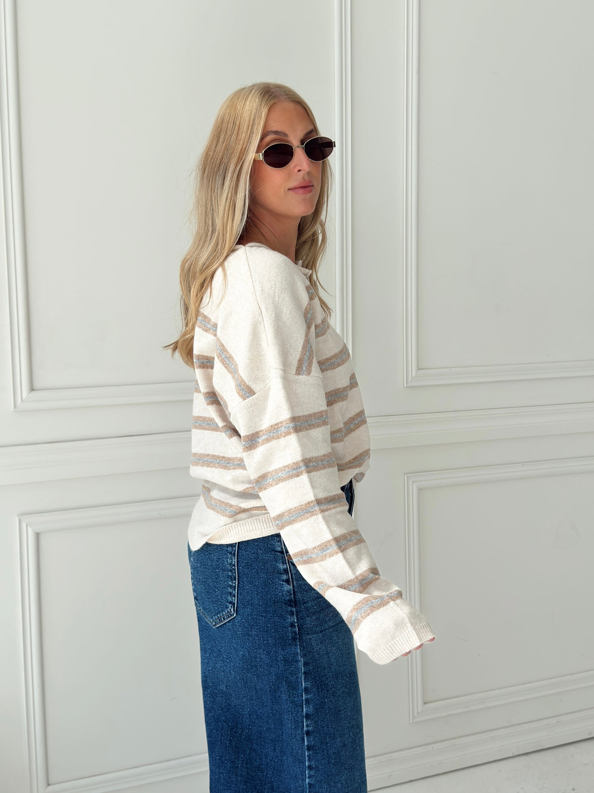 A woman with blonde hair and sunglasses wearing a cream, light blue, and beige striped long-sleeve knit top and dark blue denim jeans, standing in front of a white paneled wall.