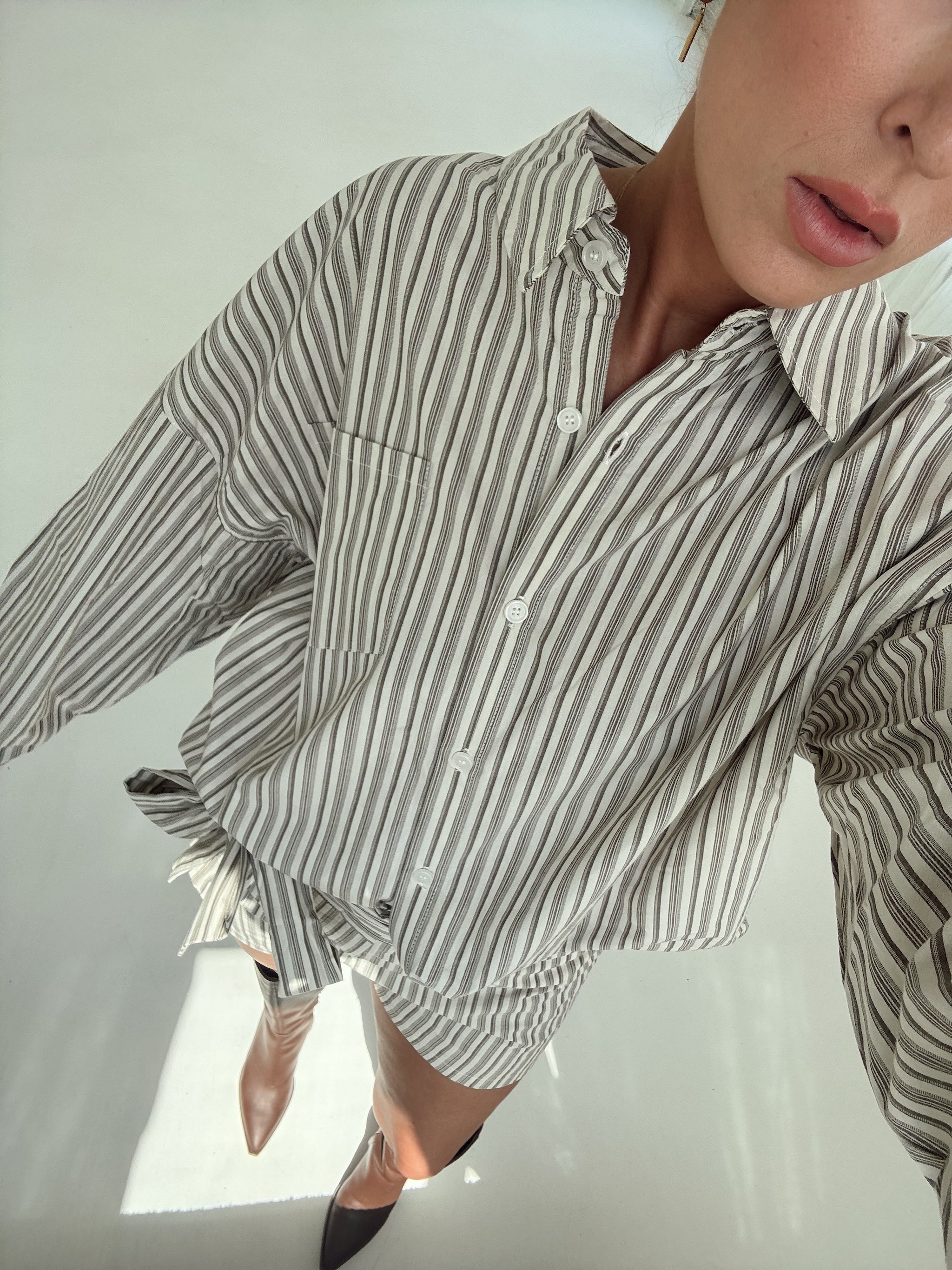 A close-up lifestyle shot of a person wearing a long-sleeve button-up shirt with vertical brown and white stripes, styled with brown pointed-toe heels against a bright, minimalist background.