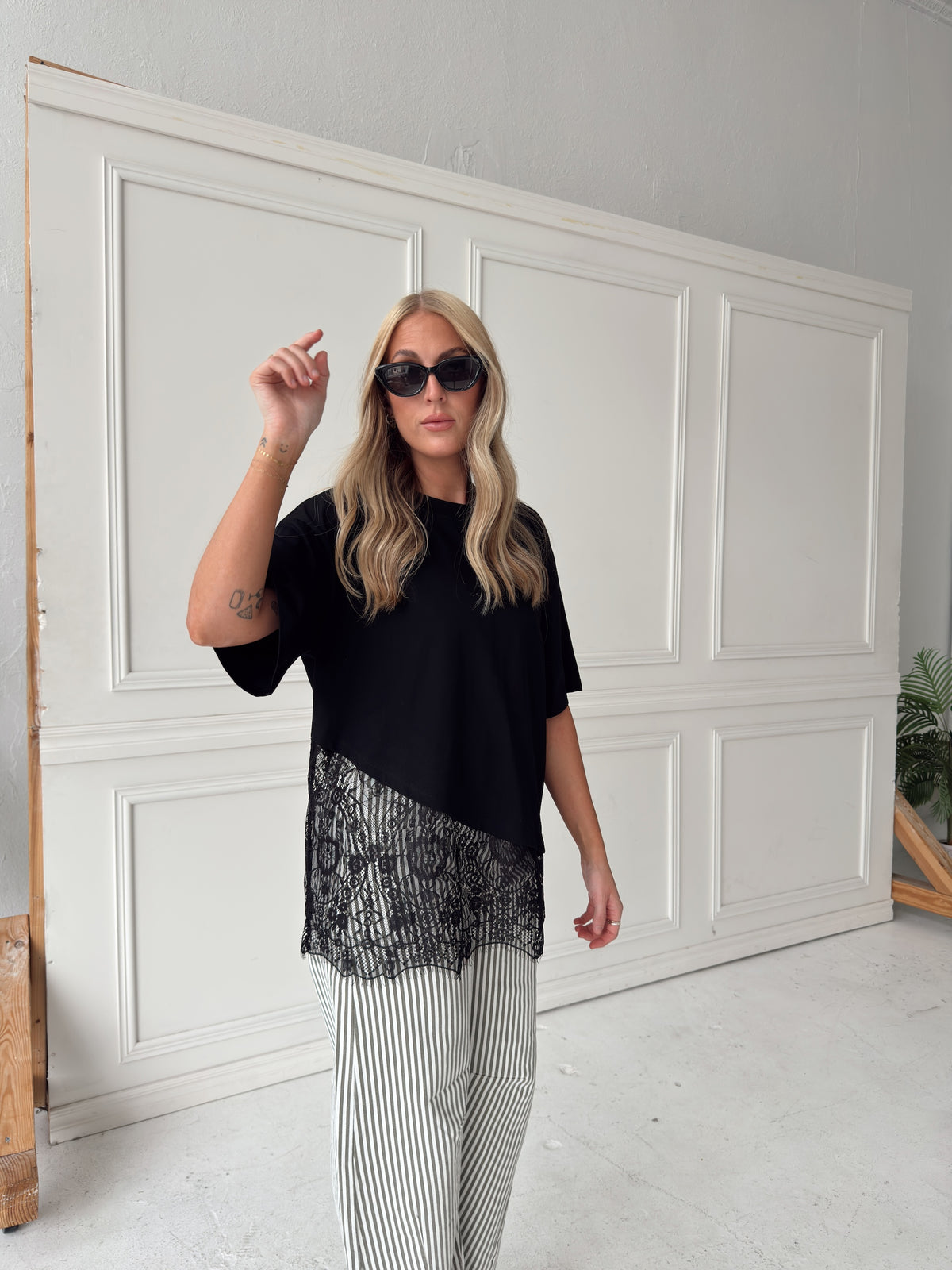 A woman modeling a black asymmetrical top with a sheer black lace hem, paired with white and olive vertically striped pants, standing in front of a white paneled studio backdrop.
