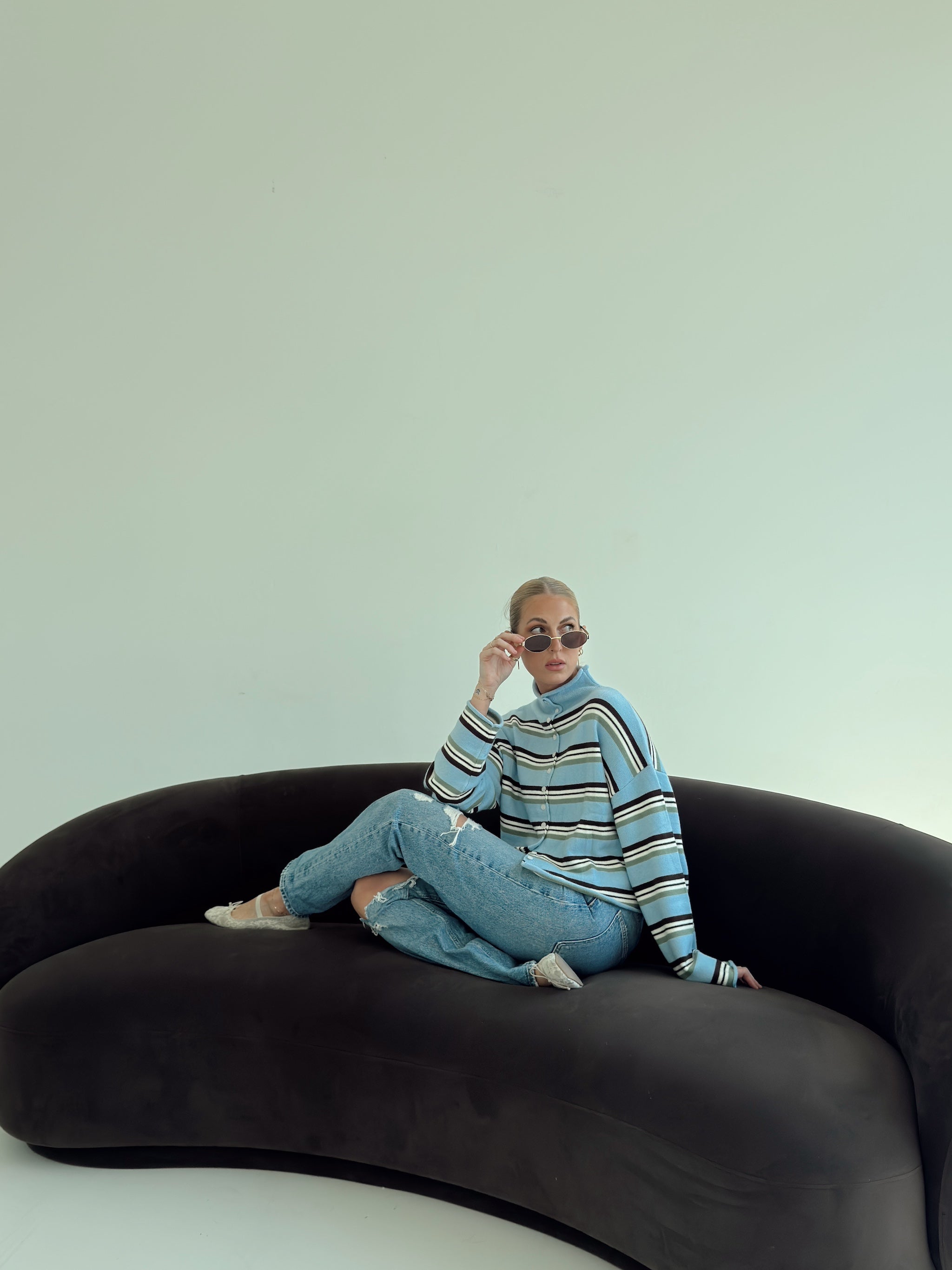 A person sitting on a curved black sofa wearing a light blue and white striped knit sweater, light blue jeans, and sunglasses against a pale mint wall.