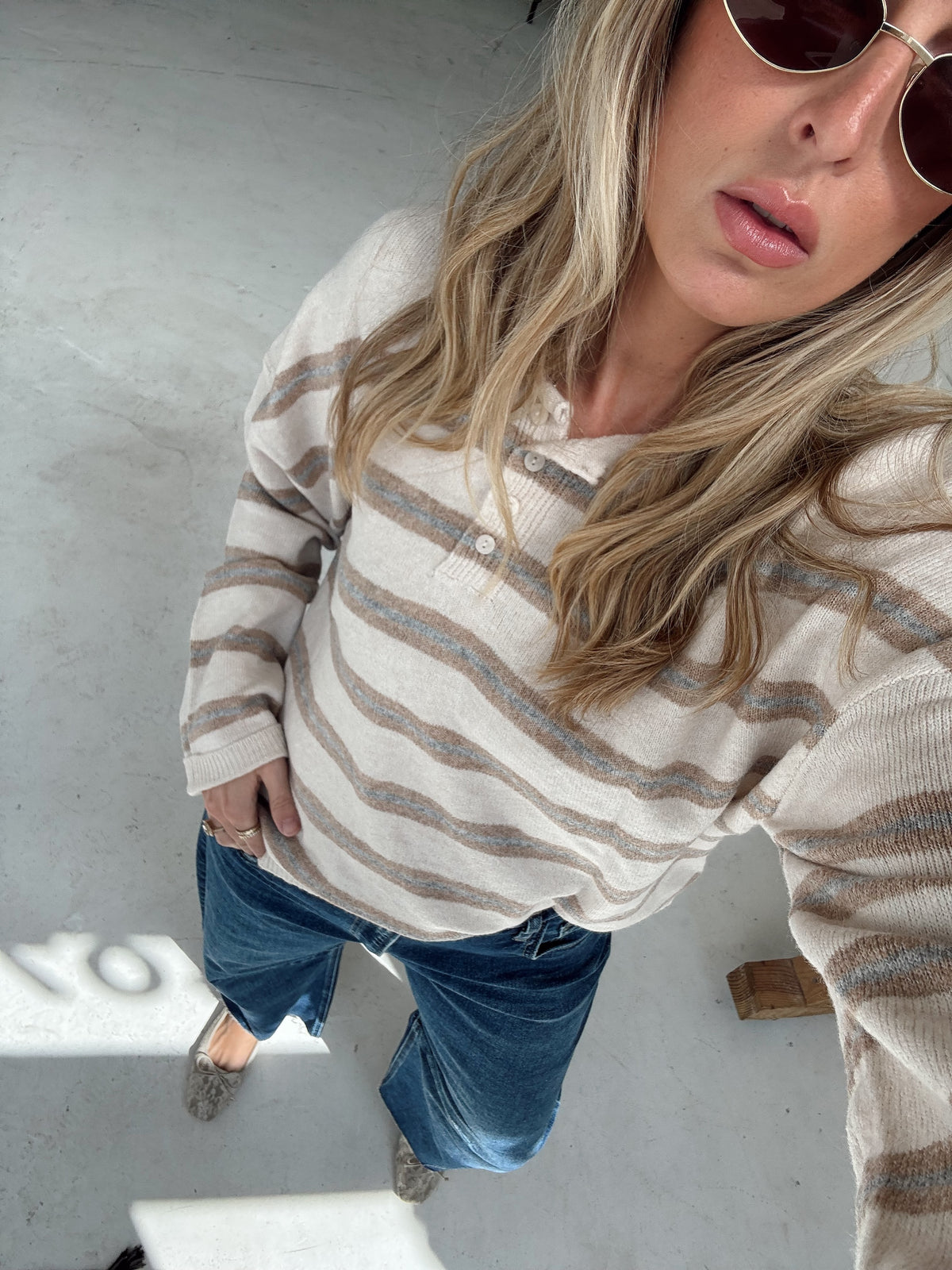 A woman wearing an oatmeal, cream, and light blue striped long-sleeve top, dark blue wide-leg jeans, and beige flats poses for a high angled selfie.