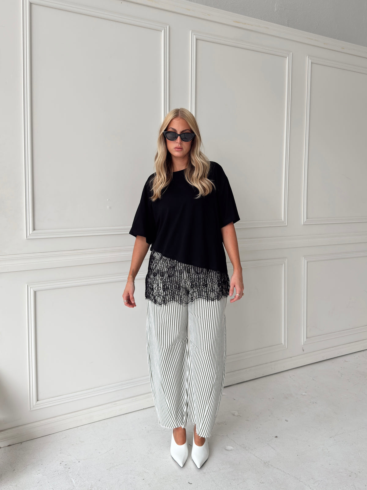 A woman modeling a pair of wide-leg barrel jeans with thin olive and white vertical stripes, paired with a black t-shirt, black sunglasses, and white pointed-toe heels, standing in front of a white paneled wall.