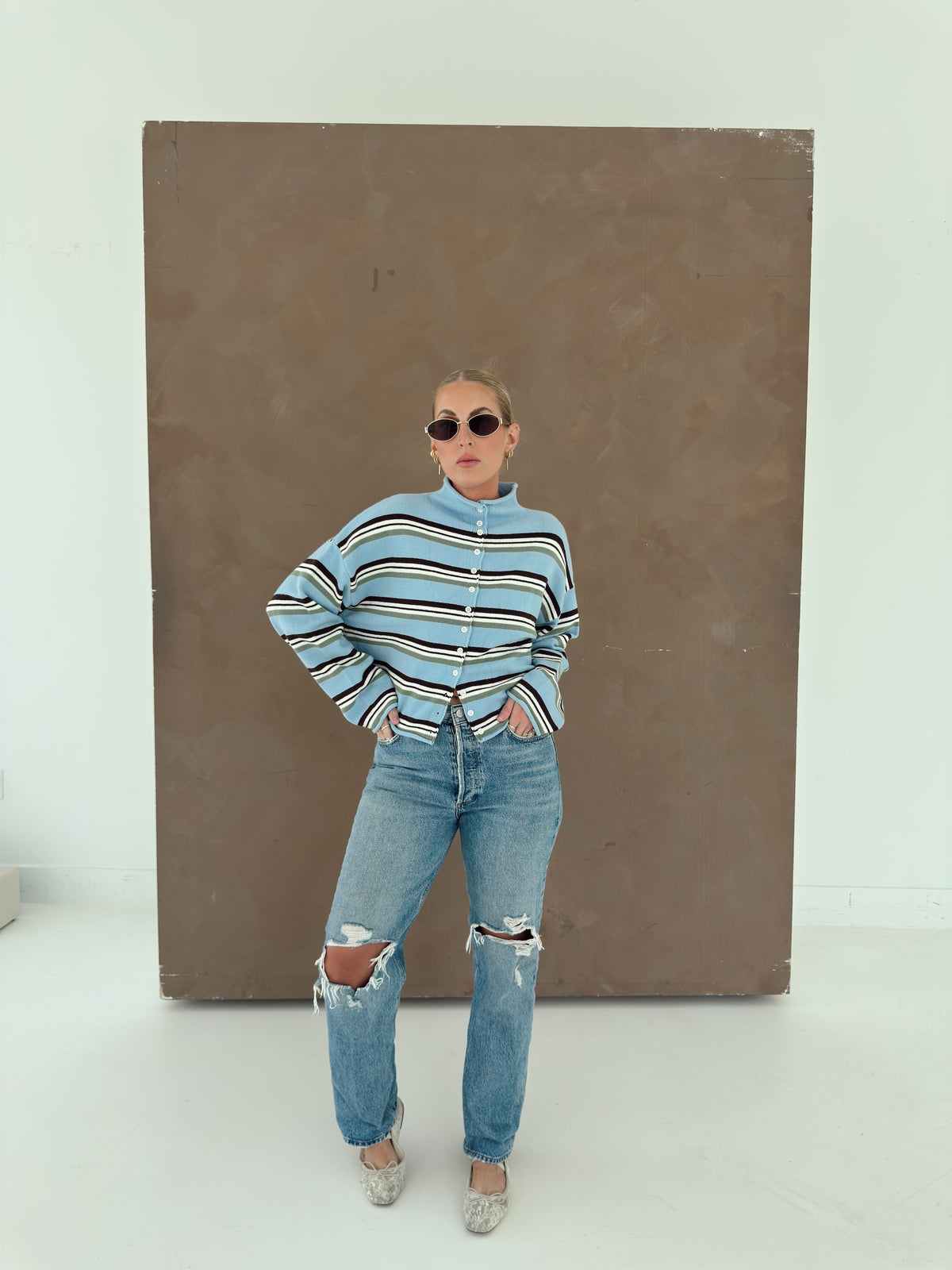 A person wearing a light blue knit cardigan with horizontal brown, white, and olive green stripes, paired with light-wash jeans and lace flats.