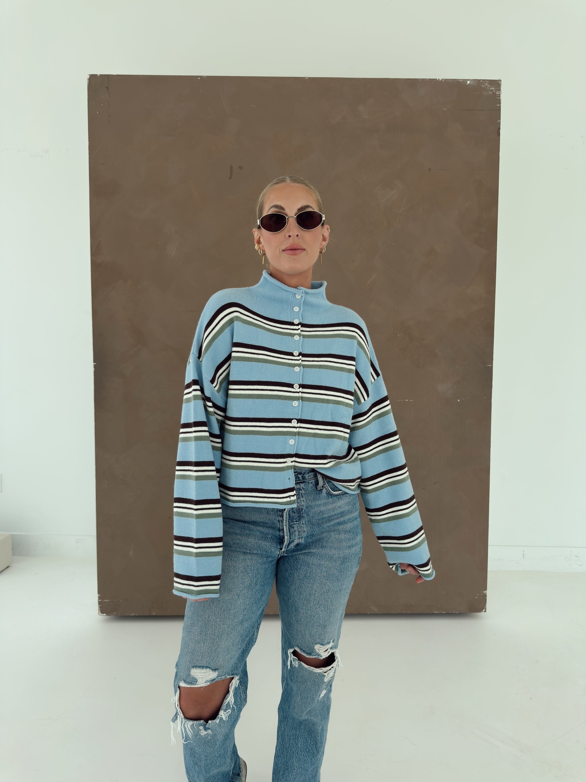 A person wearing a light blue knit cardigan with horizontal brown, white, and olive green stripes, paired with light-wash jeans and lace flats.