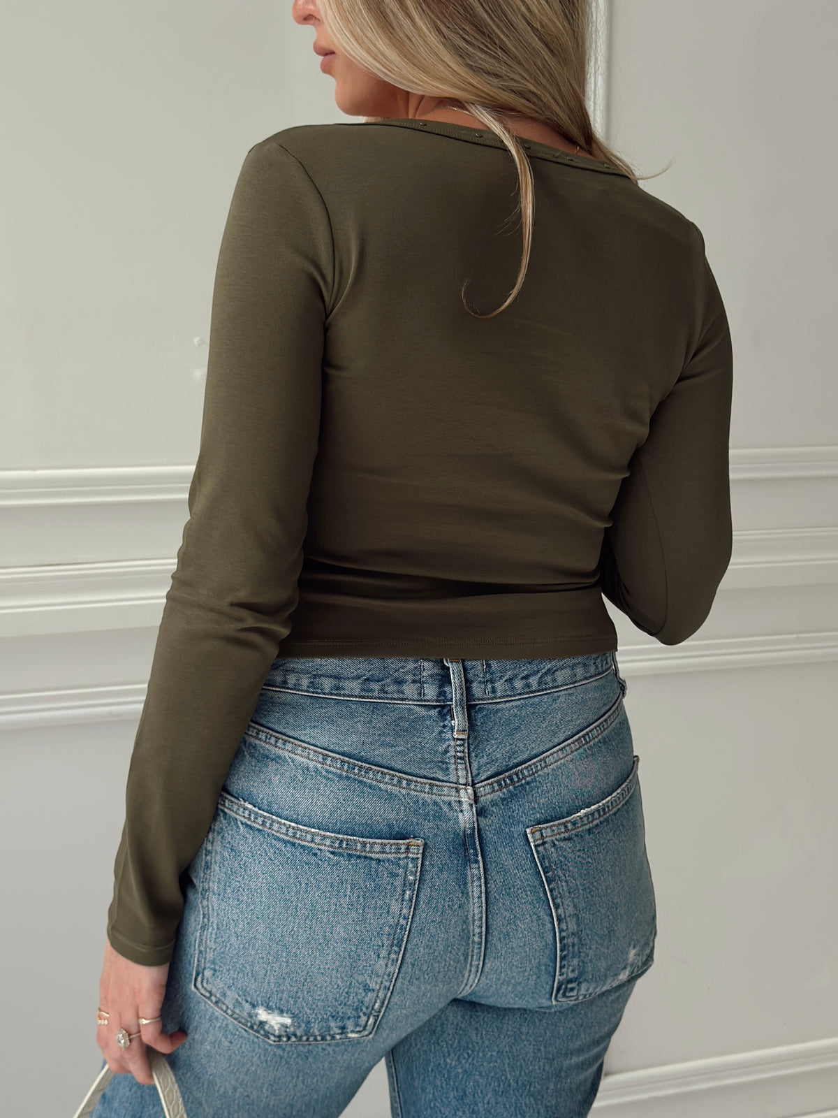 A woman shown from the back wearing a fitted long-sleeve olive green top and light blue jeans against a white paneled wall.