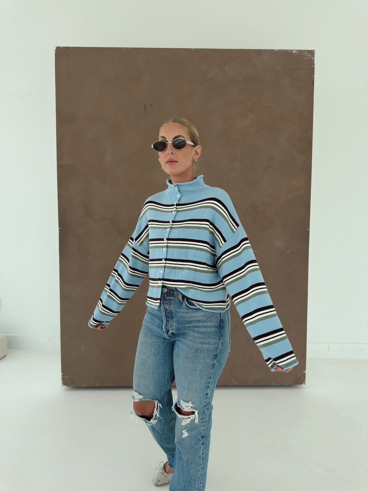A woman modeling a light blue cropped sweater with brown, white, and green horizontal stripes, paired with distressed blue jeans and sunglasses, standing in front of a large brown canvas on a white studio background.