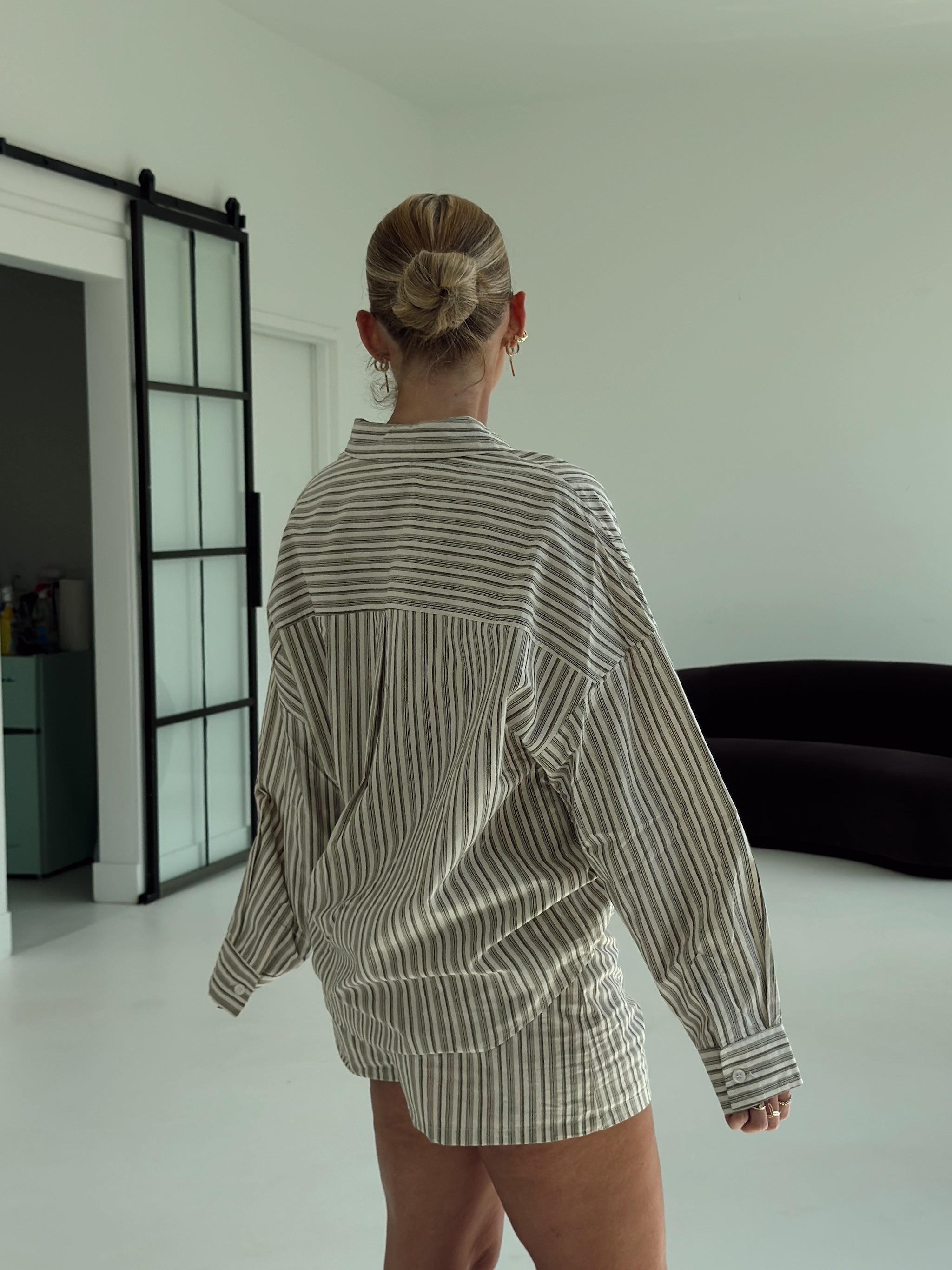 A woman viewed from behind wearing an oversized long-sleeve striped shirt in beige and brown tones, standing in a minimalist room with white walls and a black-framed glass door.