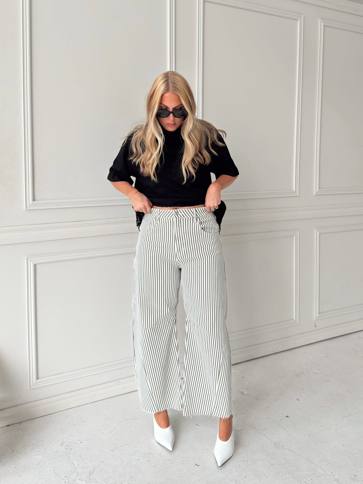 A woman modeling a pair of wide-leg barrel jeans with thin olive and white vertical stripes, paired with a black t-shirt, black sunglasses, and white pointed-toe heels, standing in front of a white paneled wall.