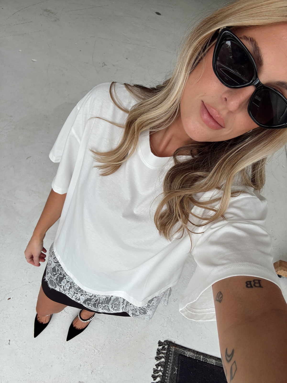 A woman wearing a white oversized short-sleeve t-shirt with a white lace hem, styled with black pointed heels and black sunglasses, taking a high-angle selfie on a concrete floor.