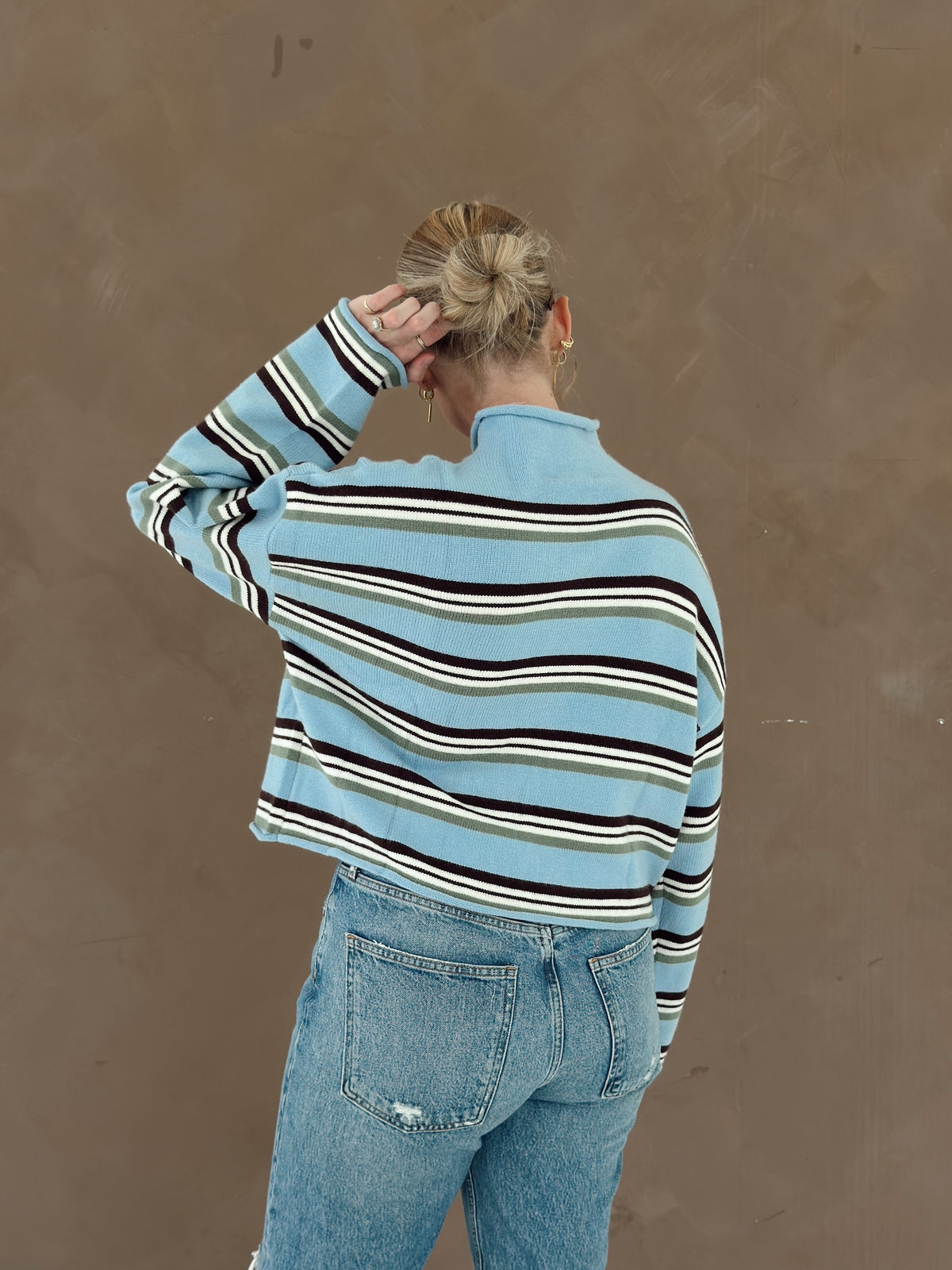 A woman shown from behind wearing a light blue cropped knit sweater with horizontal brown, white, and olive green stripes, paired with light-wash blue jeans, set against a textured brown wall.