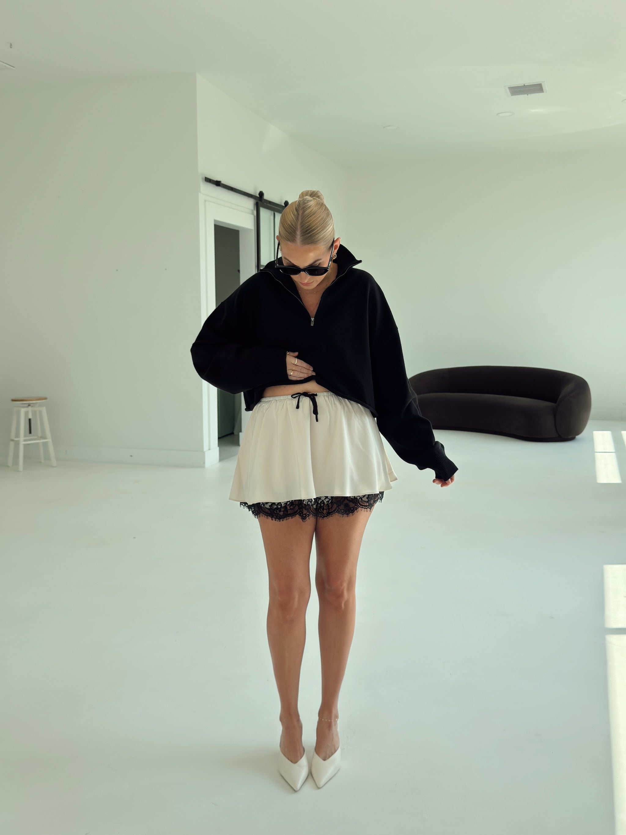 A woman stands in a bright, minimalist white room wearing a black cropped zip-up hoodie, a cream mini skirt with black lace trim, and white pointed-toe heels.