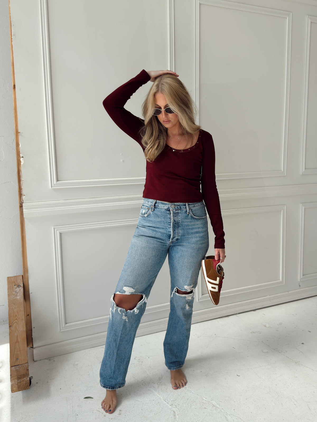 A woman stands barefoot in a bright room with white paneled walls, wearing a fitted burgundy long-sleeve top, light-wash ripped blue jeans, and holding brown shoes.