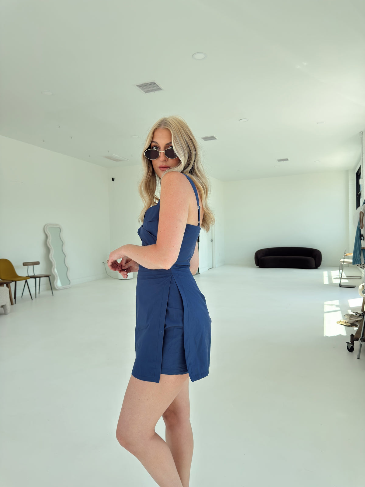 A woman modeling a sleeveless blue romper in a bright, minimalist white studio space.