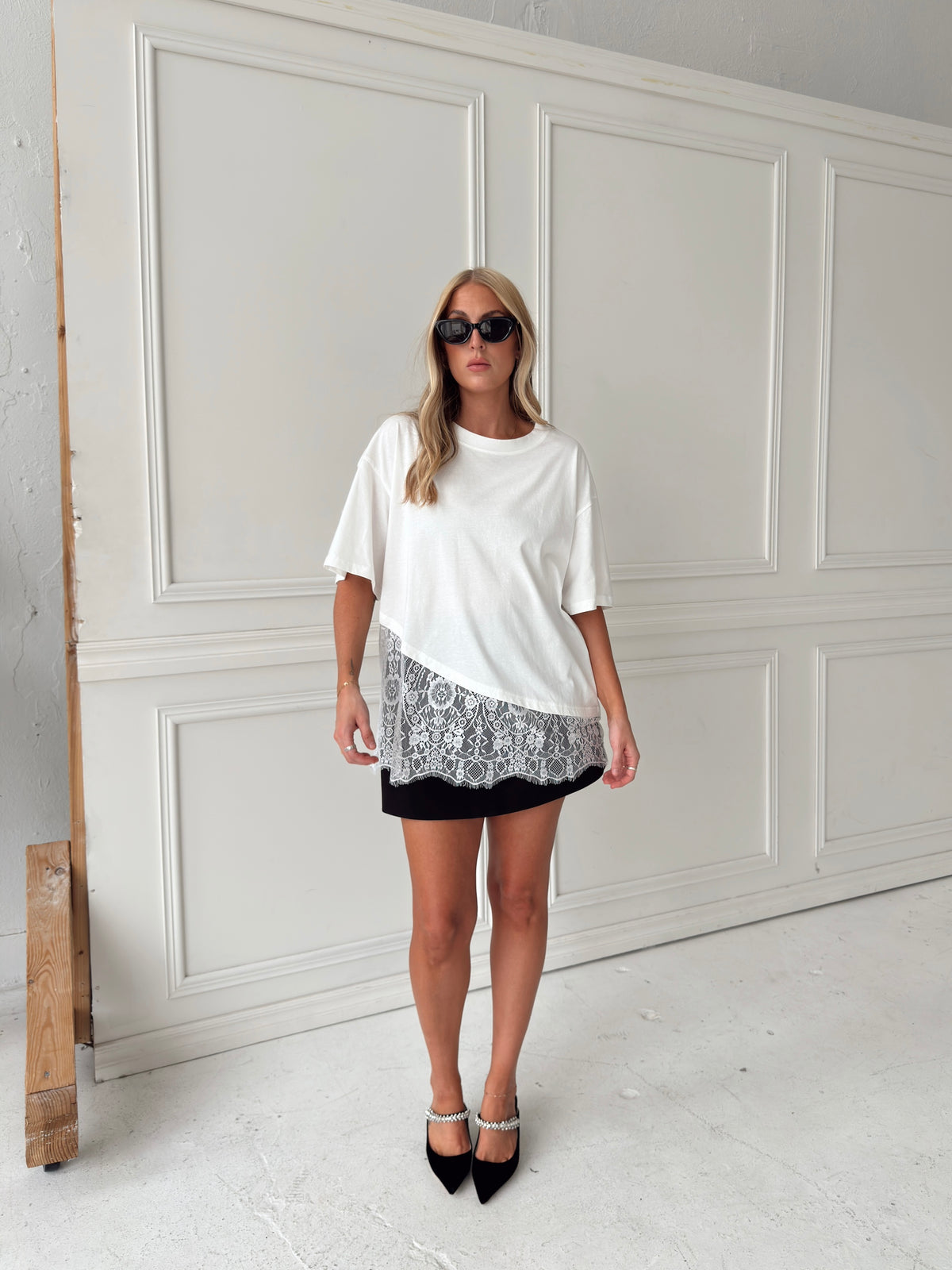 A woman modeling a white short-sleeve top with white lace hem and a black skort, paired with black pointed-toe heels, standing against a white paneled wall.